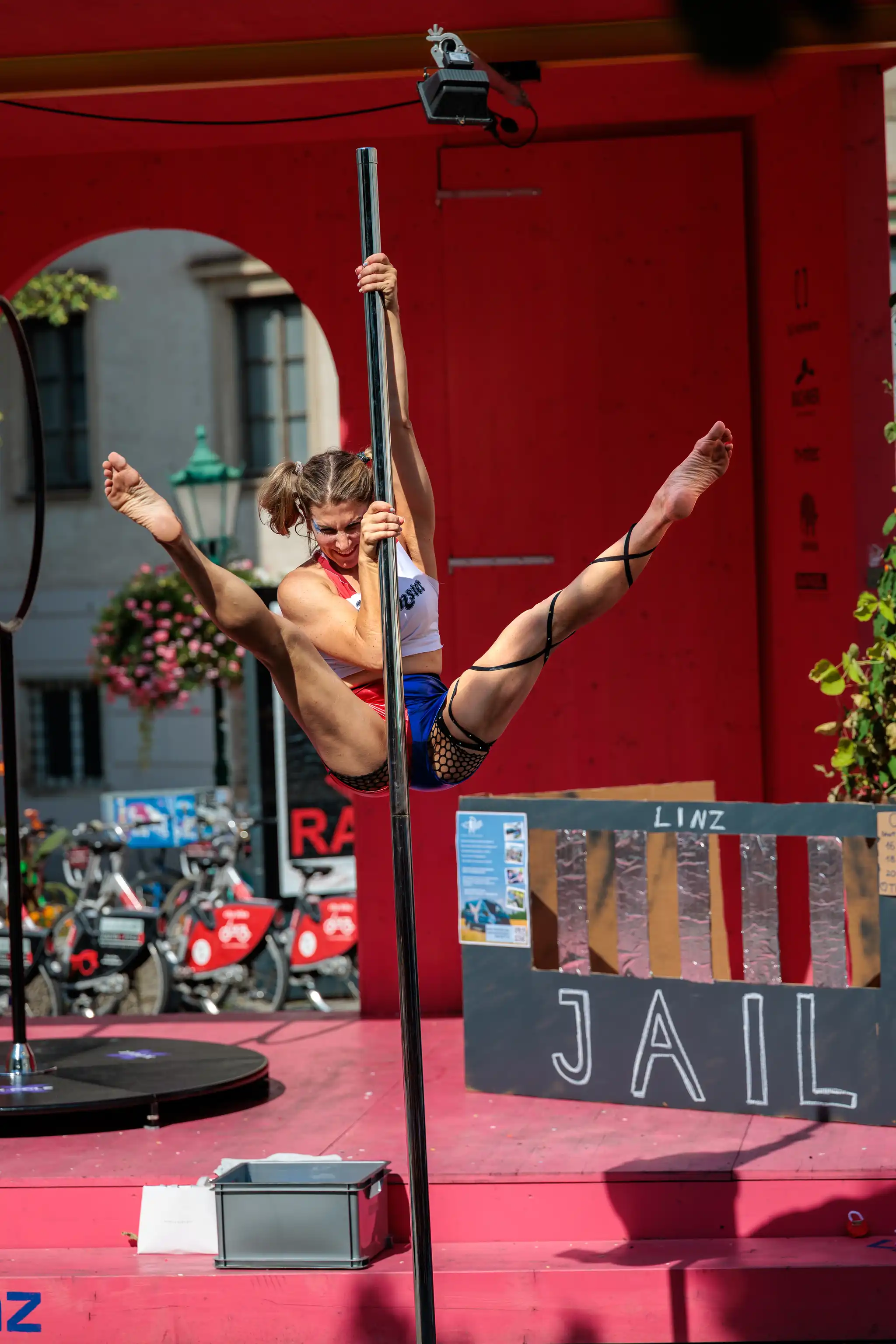Athletische Frau beim Poledance im Freien, die eine anspruchsvolle Spagat-Pose an der Stange hält. Sie trägt Sportbekleidung und Fischnetzstrümpfe. Im Hintergrund sind eine rote Bühne, Gebäude, Fahrräder und ein Schild mit der Aufschrift „LINZ“ und „JAIL“ zu sehen. Der Auftritt war im Rahmen des Pflasterspektakels 2024