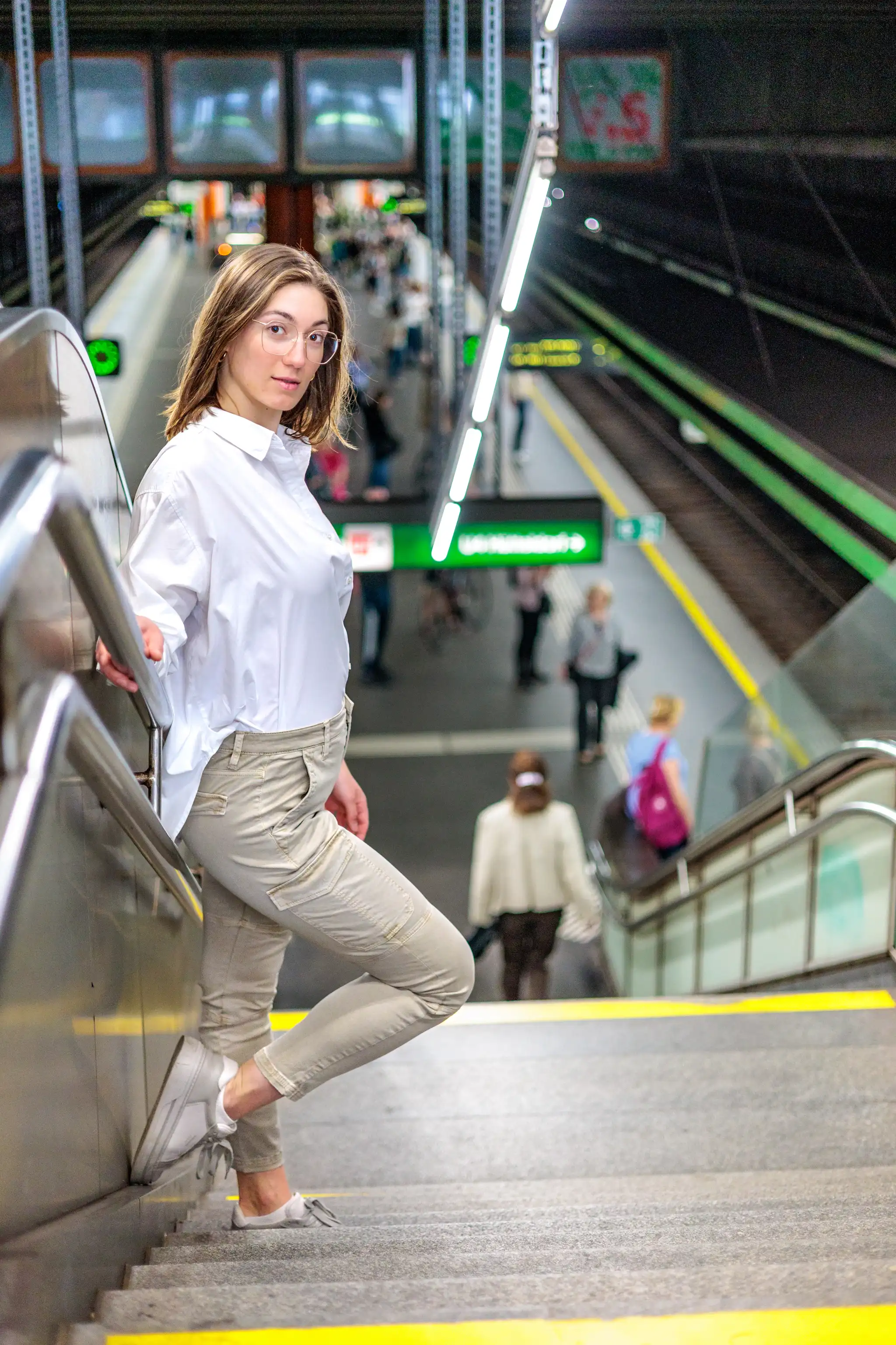 Junge Frau mit Brille in weißer Bluse und beiger Cargohose posiert auf Treppe einer Wiener U-Bahn-Station
