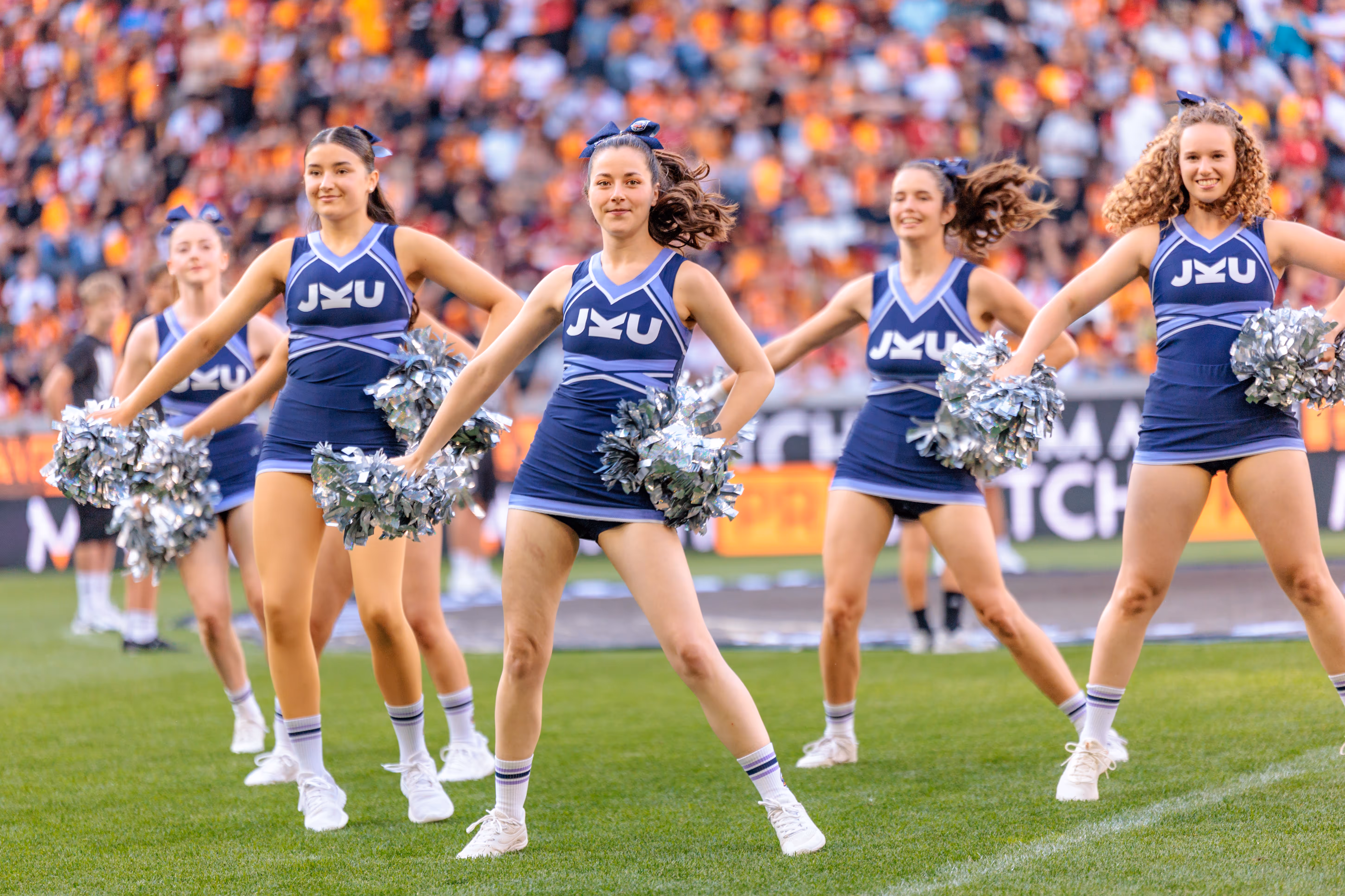Eine Gruppe junger Cheerleaderinnen in blauen JKU-Uniformen mit silbernen Pompons tritt vor einer Stadionkulisse auf