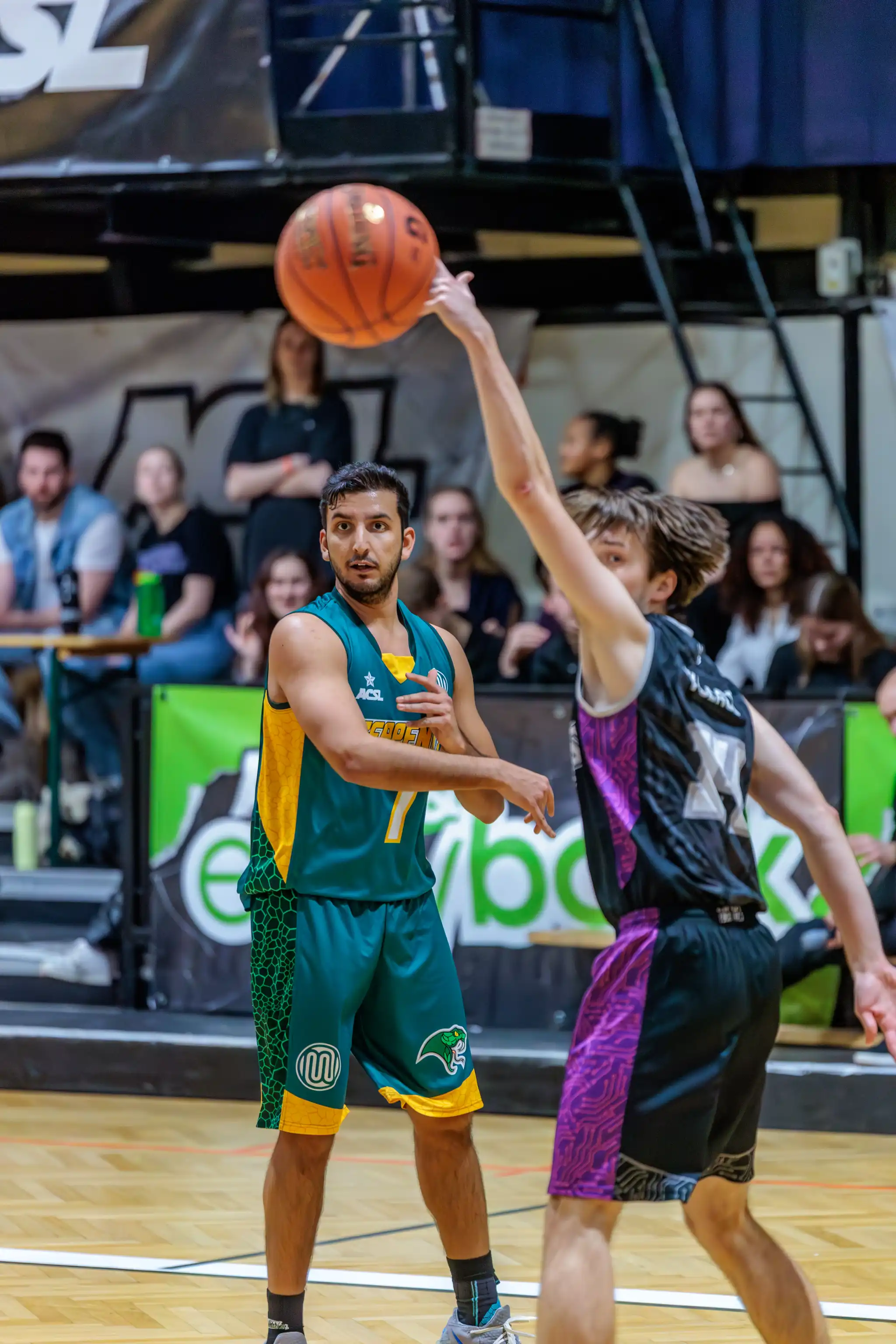 Zwei männliche Basketballspieler auf einem Hallenplatz: Einer wirft den Ball, der andere, in grün-gelbem Trikot mit Nr. 7, beobachtet. Im Hintergrund sind Zuschauer zu sehen