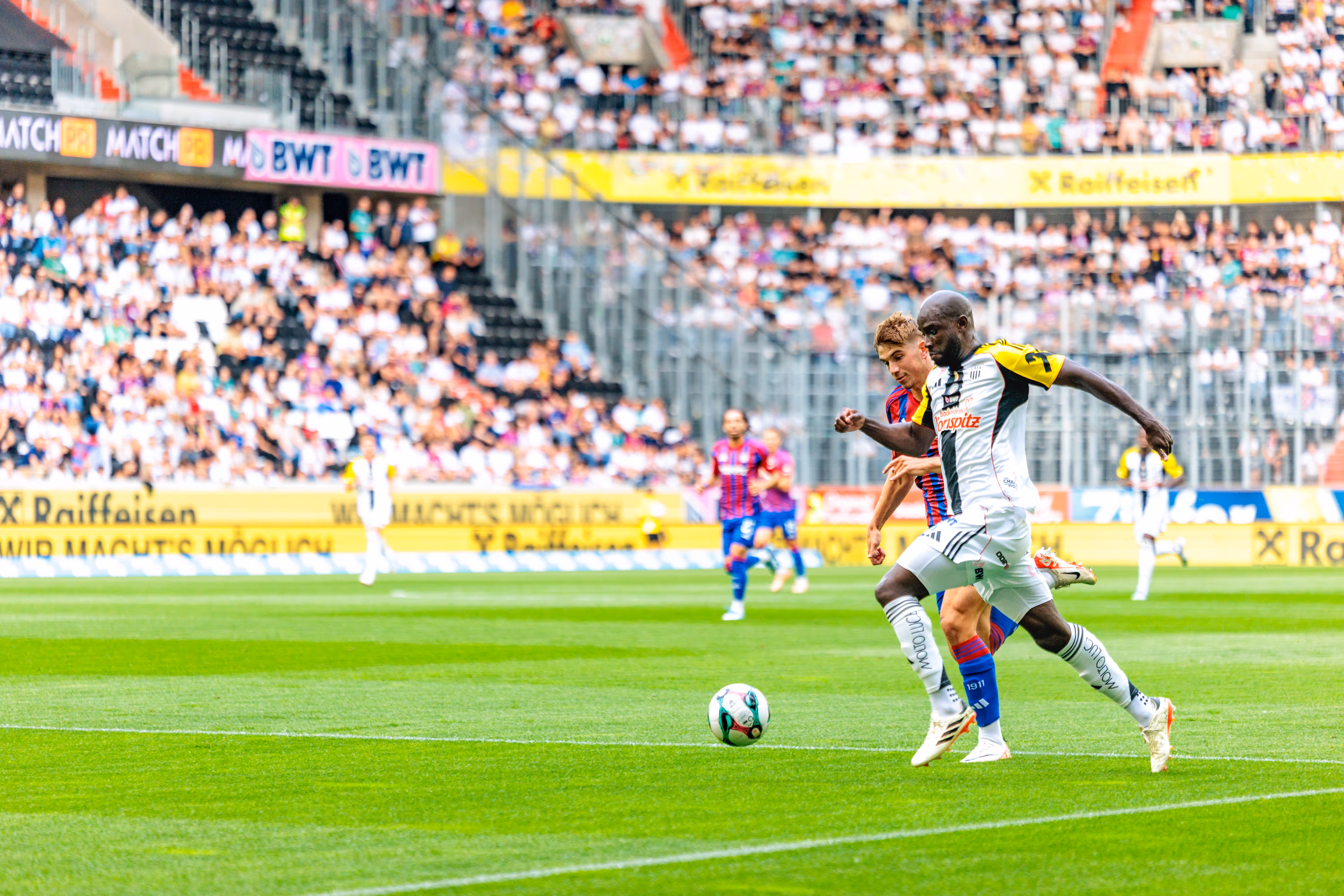 Zwei Fußballspieler im Duell um den Ball während eines Spiels in der Raiffeisen Arena, umgeben von Fans