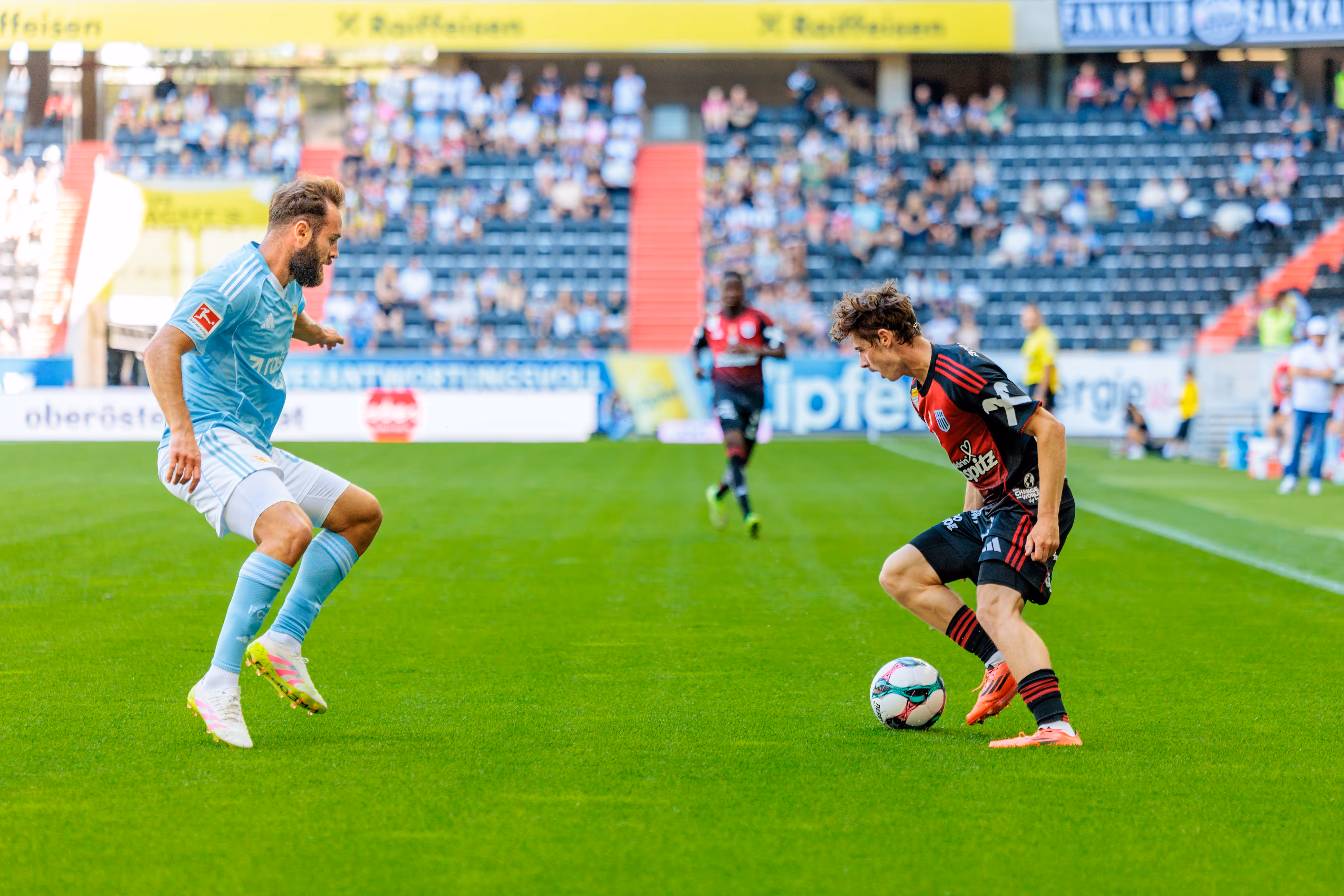 Ein Fußballspieler in hellem Trikot blickt auf seinen Gegner in dunklerem Trikot, der den Ball am Fuß hat, während eines Spiels vor Stadionpublikum - 2025/07/19