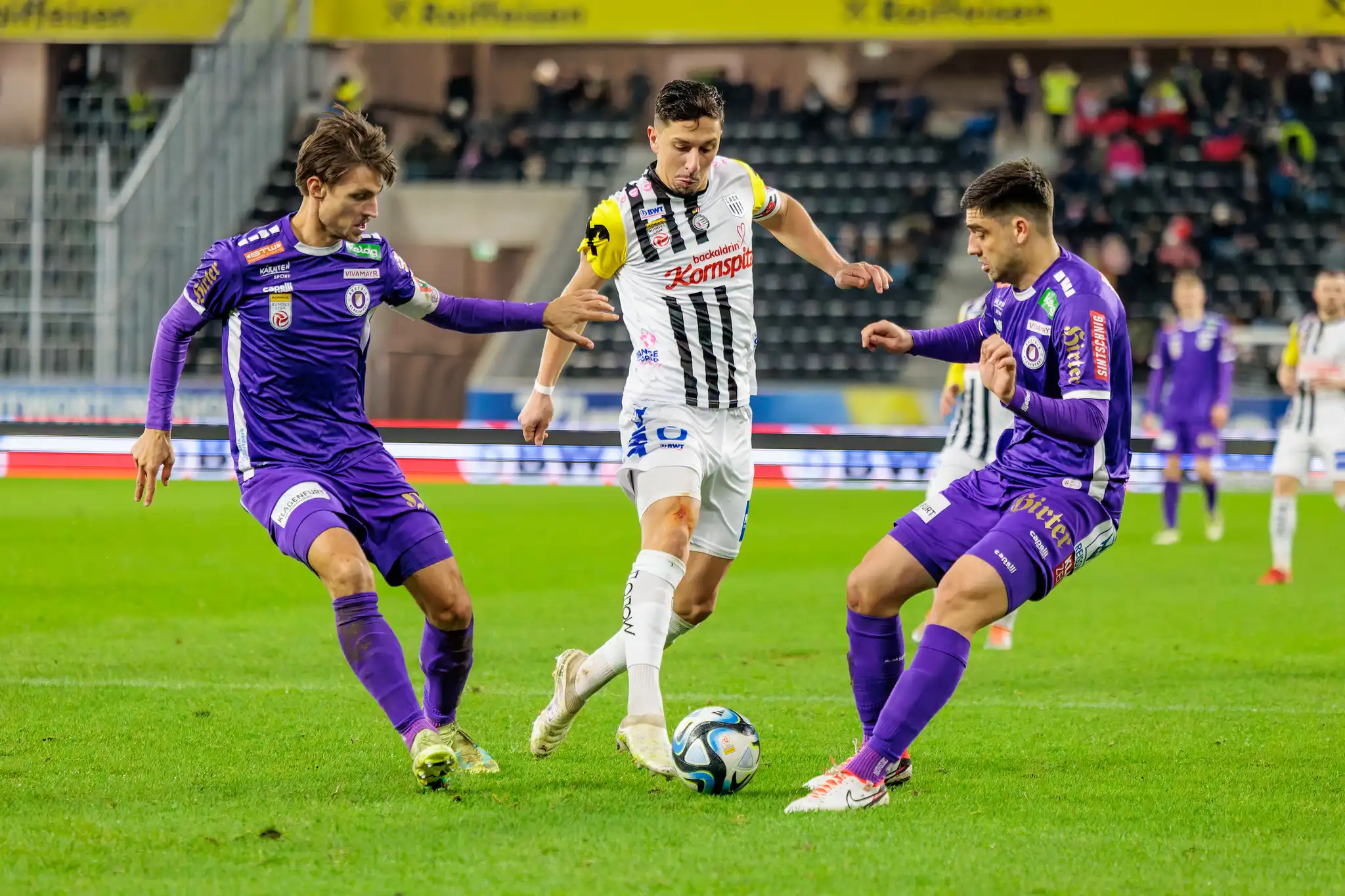 Ein Spieler im schwarz-weiß gestreiften Trikot im Zweikampf mit zwei Gegnern in lilafarbenen Trikots um den Fußball während einer Bundesliga-Partie
