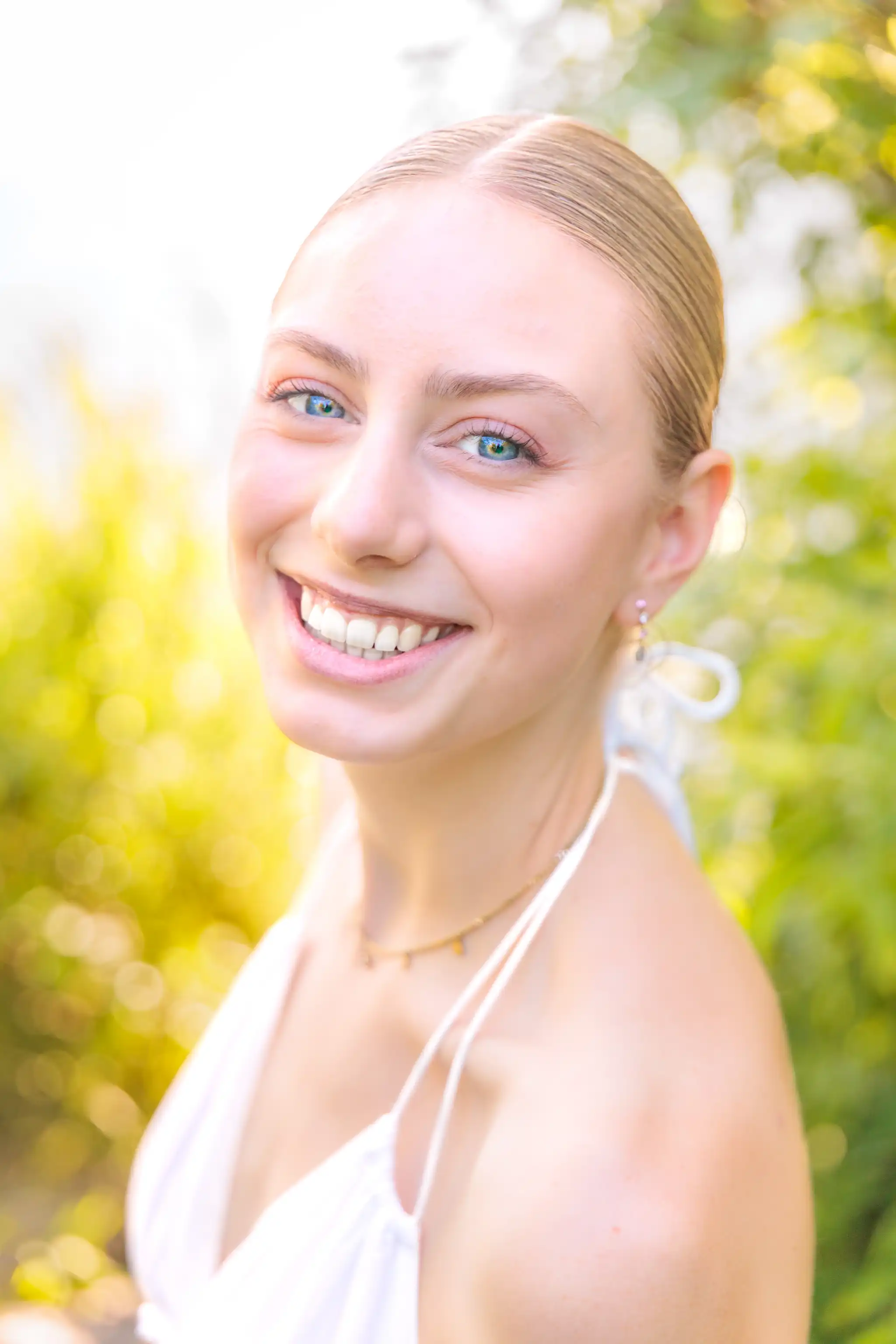 Porträt einer lächelnden jungen Frau mit blonden Haaren, blauen Augen und einem weißen Kleid im Freien mit verschwommenem Hintergrund aus grünen Blättern und goldenem Licht. 