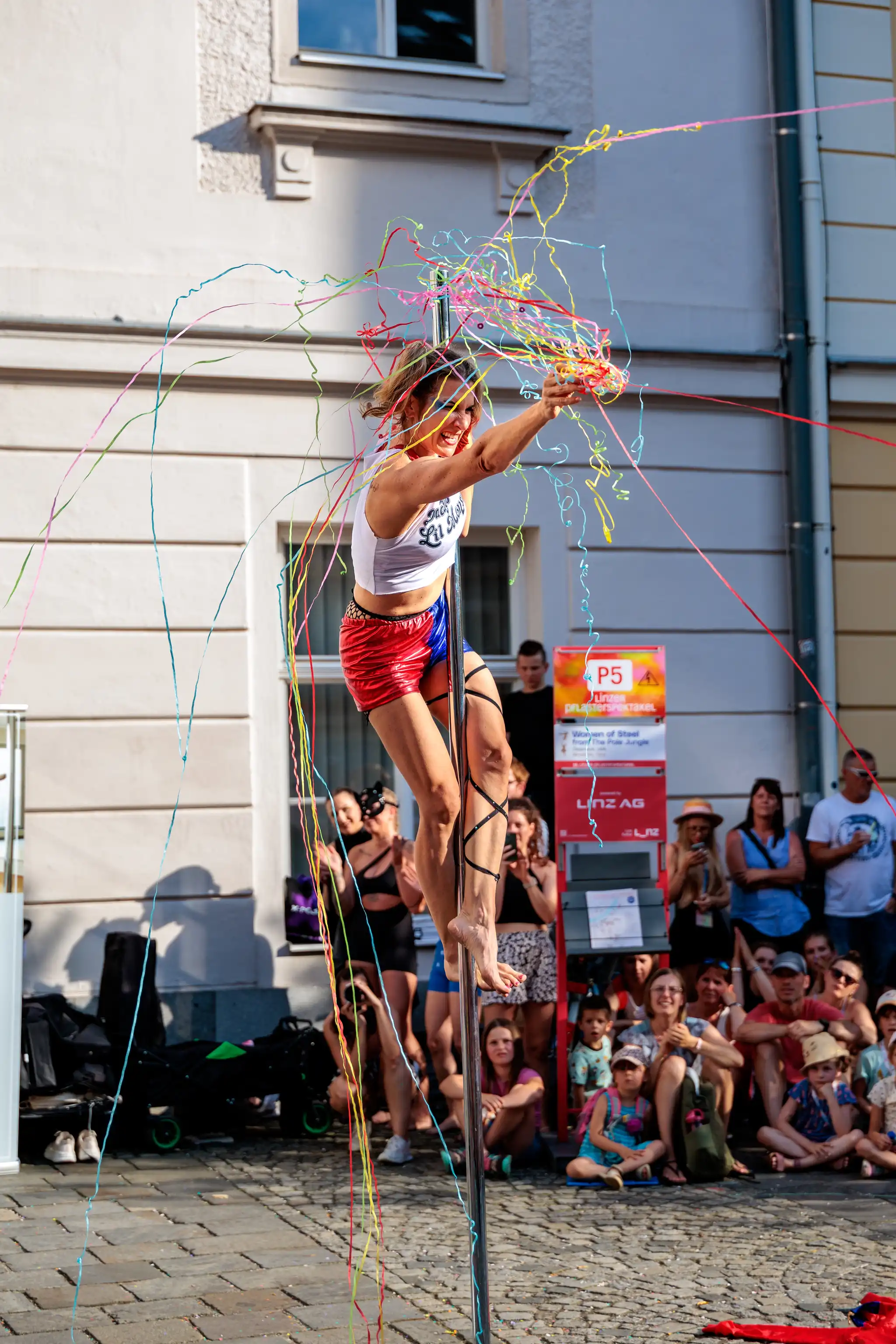Eine energische Pole-Tänzerin in rot-blauen Shorts und weißem Top wirft buntes Konfetti während einer Outdoor-Performance vor dem Publikum des Pflasterspektakels in Linz