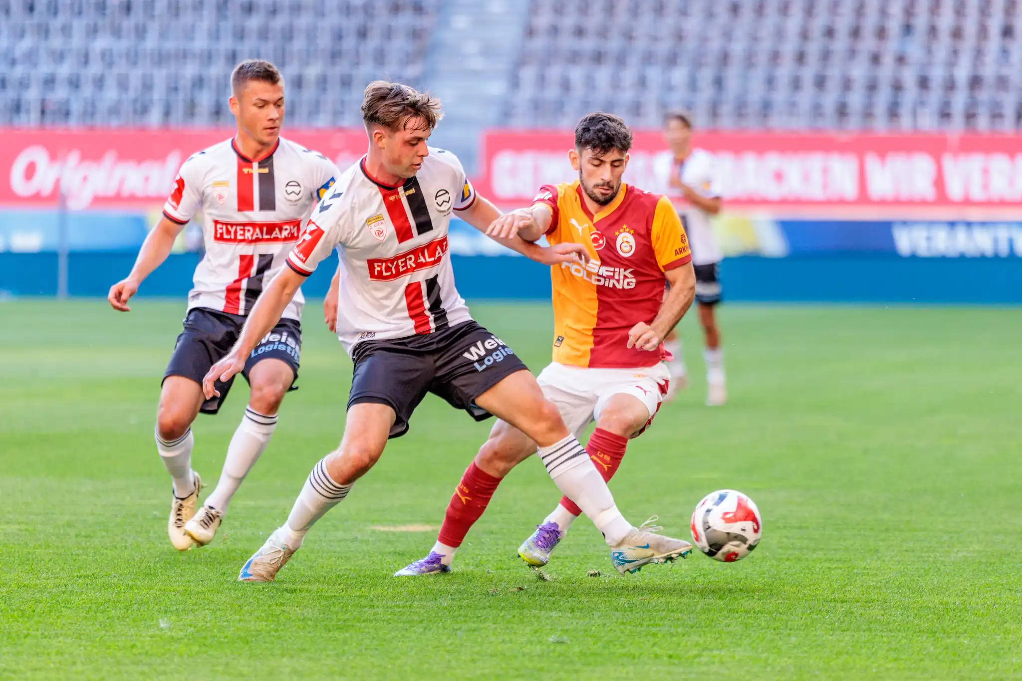 Zwei Fußballspieler in Trikots mit weiß-rot-schwarzen beziehungsweise rot-orangen Streifen liefern sich einen Zweikampf um den Ball 