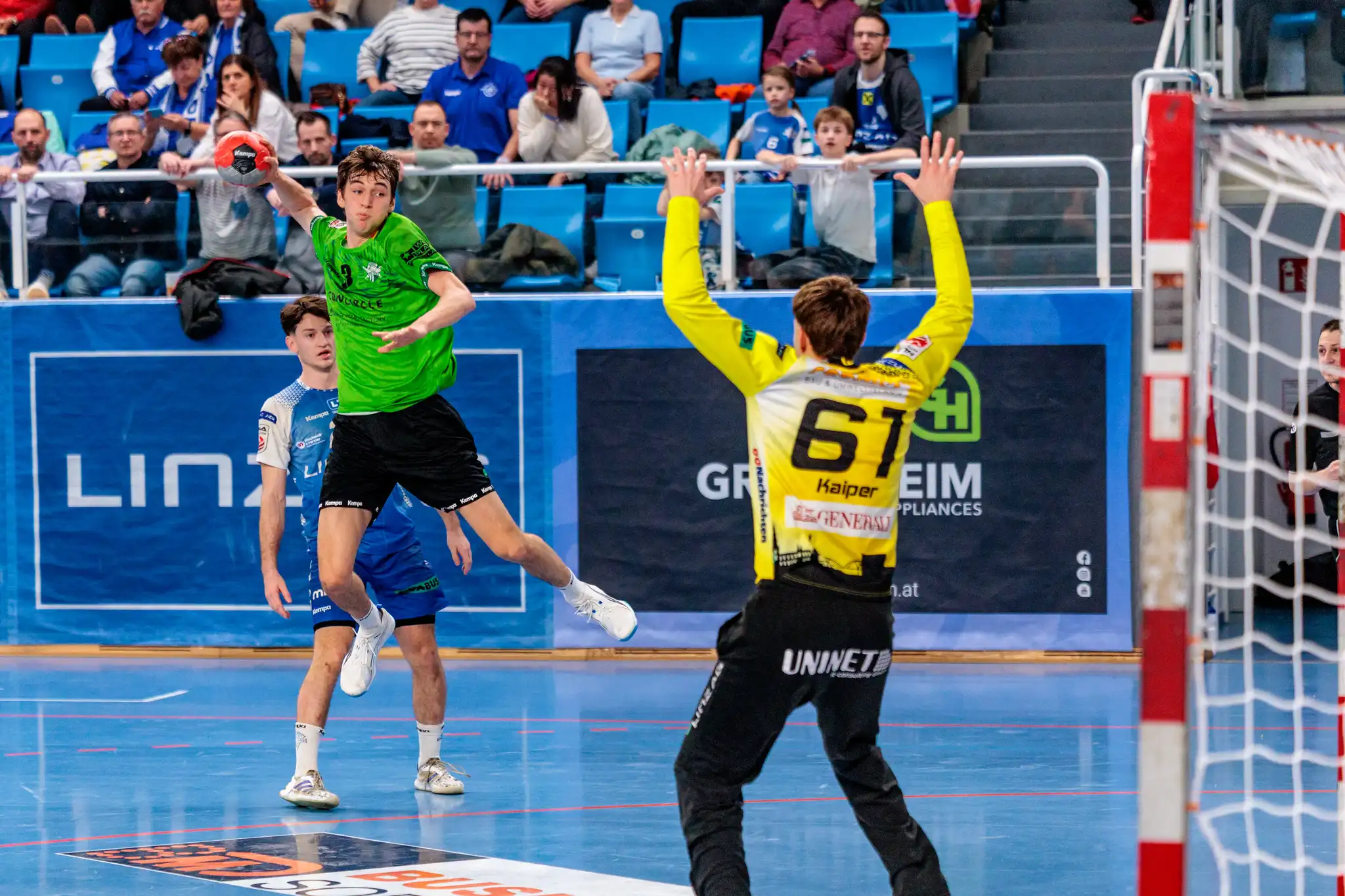 Handballspiel: Spieler in grünem Trikot beim Sprungwurf, Torwart in Gelb pariert. Dynamische Szene mit Zuschauern in Sporthalle