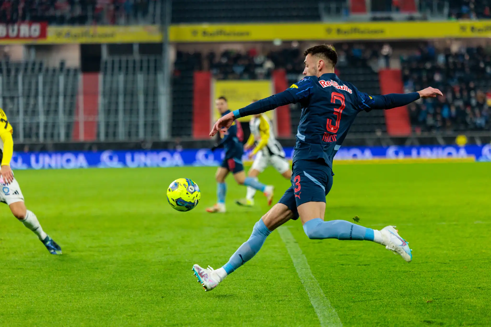 Dynamische Fußballszene: Spieler in Red Bull Trikot (Nr. 3) tritt gelb-grünen Ball auf grünem Stadionrasen