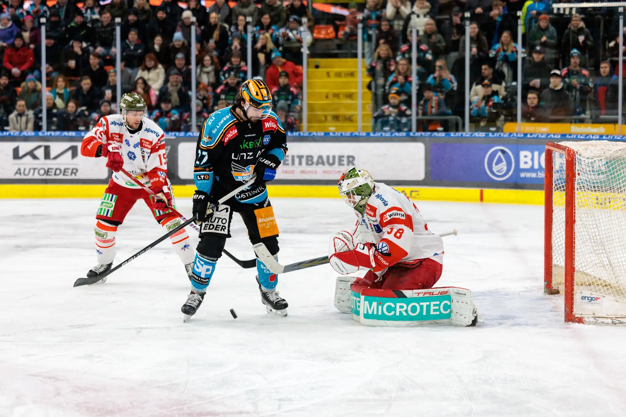 Eishockey-Spielszene mit Spielern und Torhüter in Aktion vor dem Tor, Puck auf dem Eis und Fans im Stadion der Black Wings Linz
