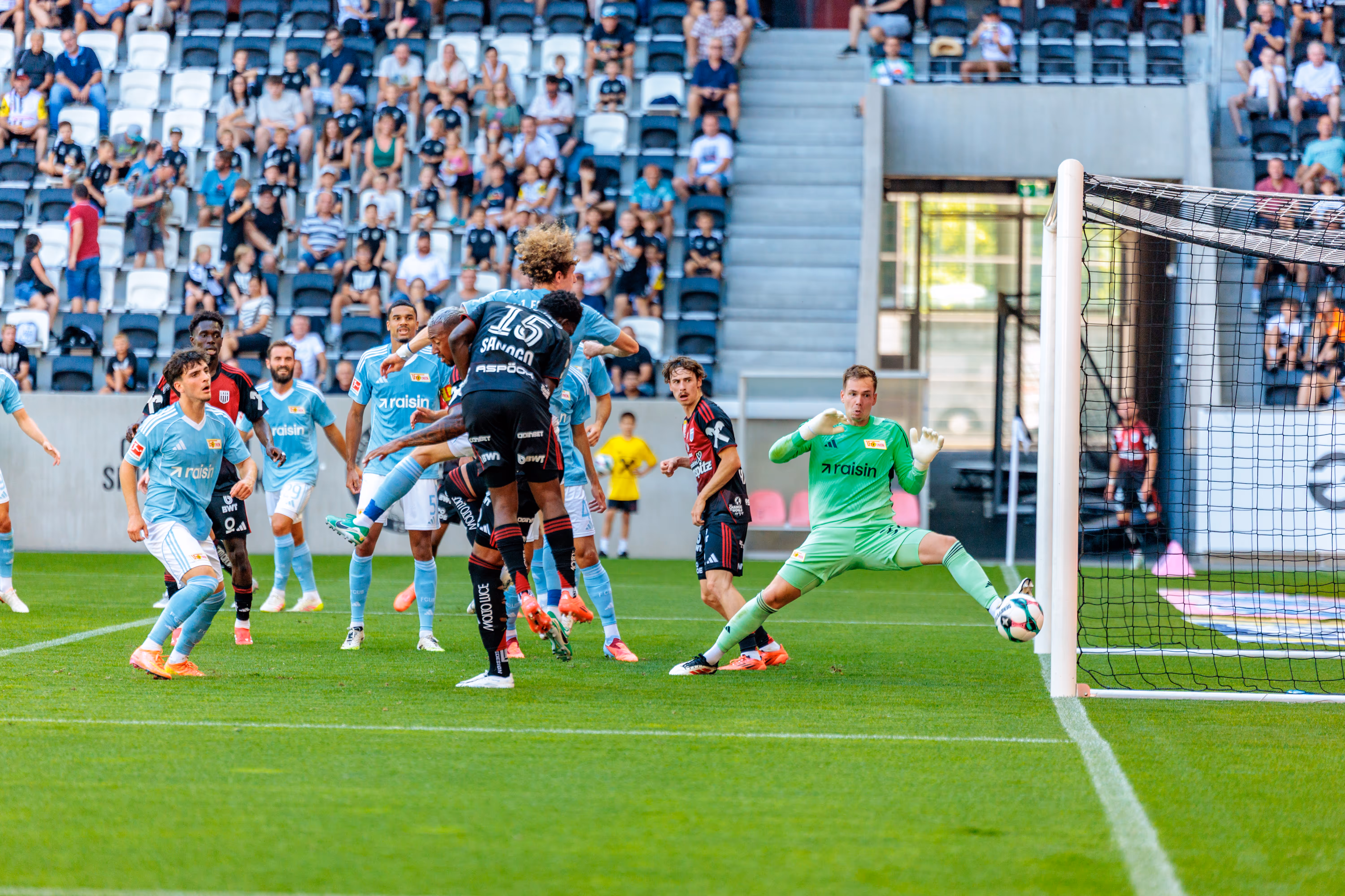 Ein grüner Torhüter hält in der Raiffeisen Arena im Sprung einen Ball vom Tor fern, umgeben von Spielern in hellblauen und dunklen Trikots