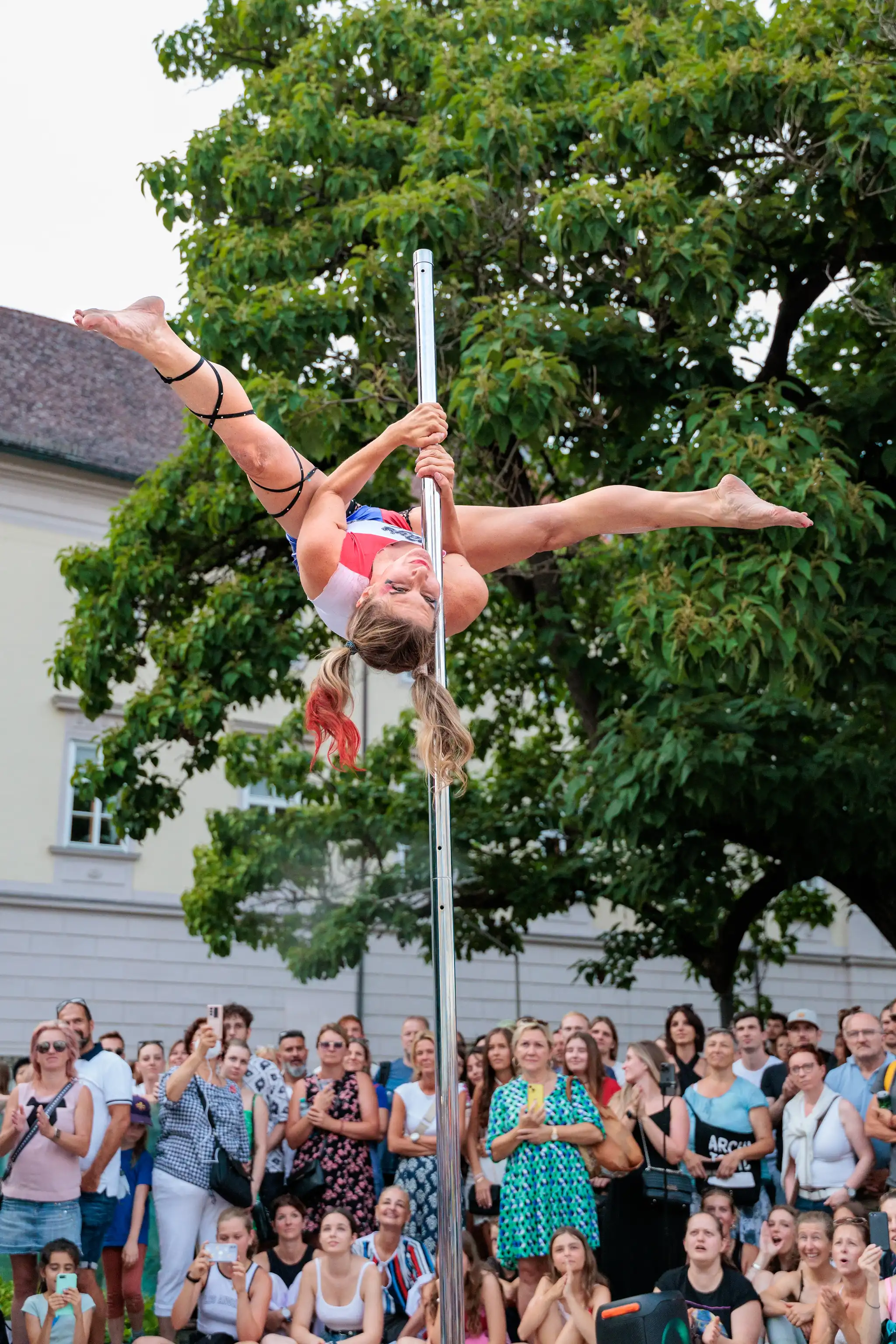 Eine Frau in Poledance-Sportkleidung vollführt kopfüber eine Spagat-Pose an einer Stange bei einer Outdoor-Vorstellung vor dem Linzer Landhaus während des Pflasterspektakels 2024