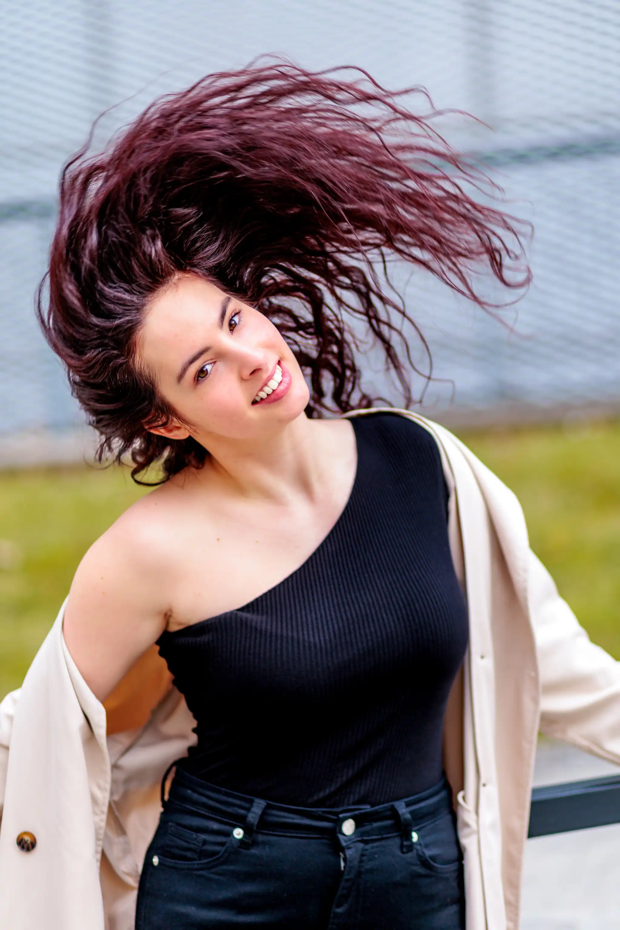 Porträt einer lächelnden jungen Frau, Nicole, mit hochgeworfenem dunklem, welligem Haar, trägt schwarzes One-Shoulder-Top und beigen Mantel bei einem Outdoor-Shooting in Linz