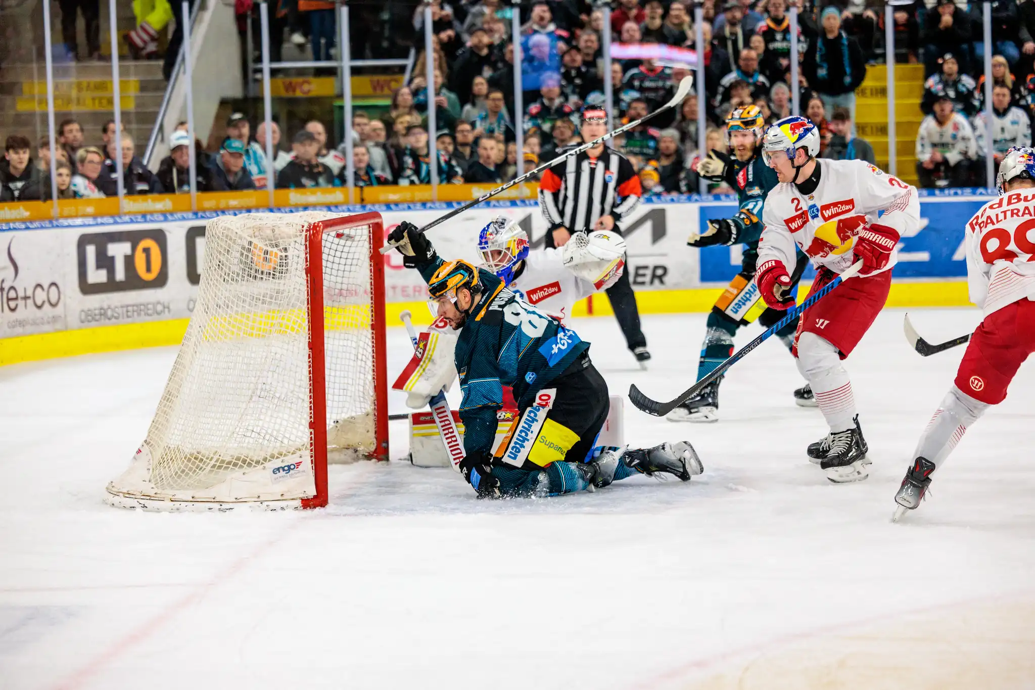 Ein Eishockey-Torwart wehrt während eines intensiven Spielzugs am Tor den Puck ab, umringt von Spielern beider Mannschaften