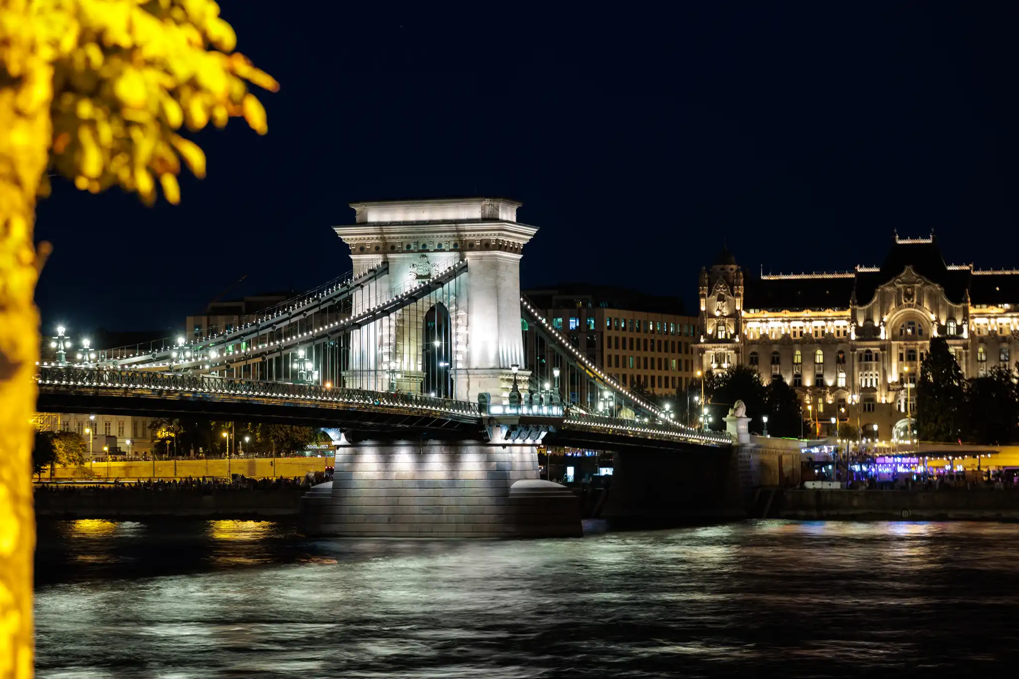 Nachtaufnahme der beleuchteten Kettenbrücke über die Donau in Budapest, Ungarn, mit Stadtansicht und Wasserspiegelungen.