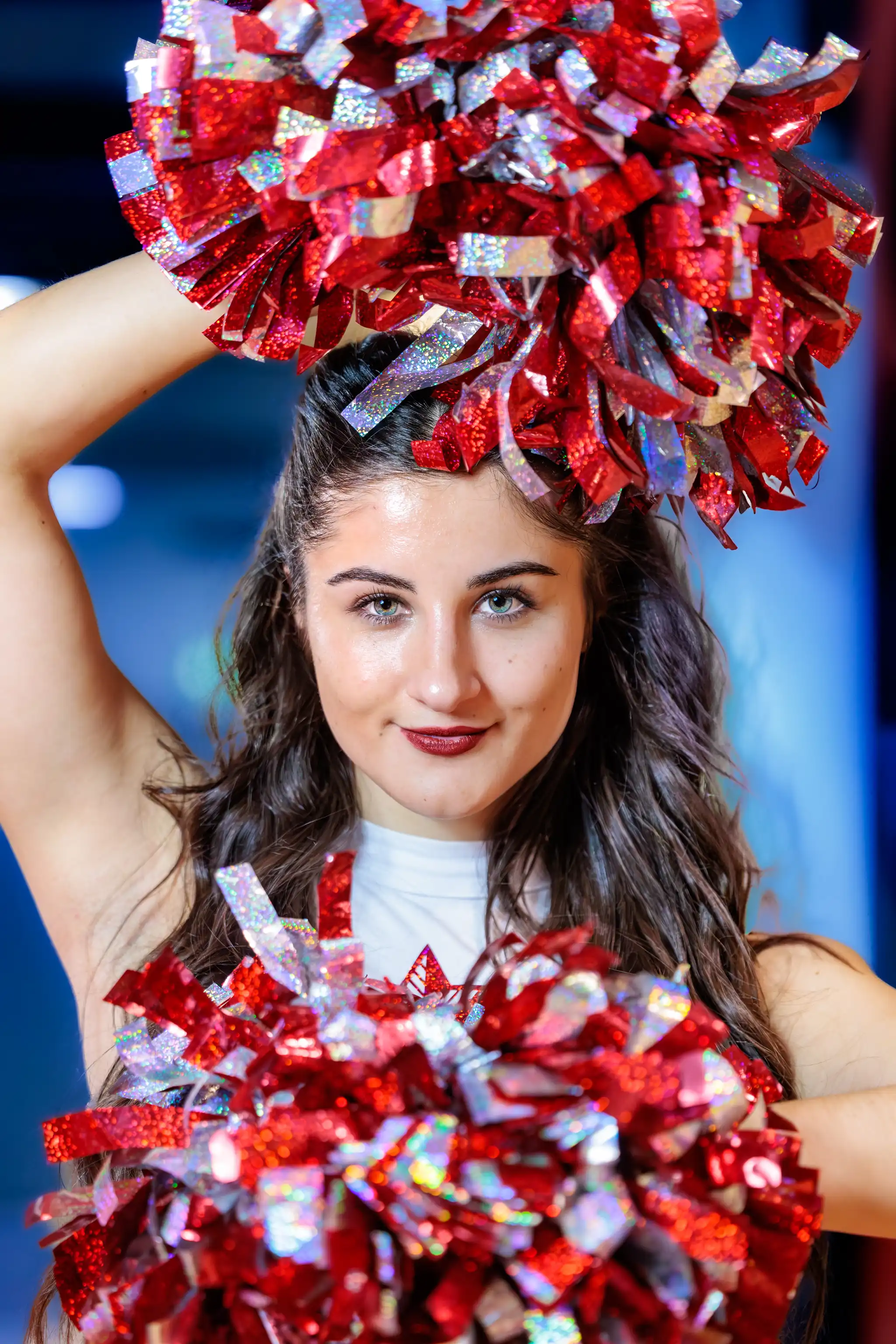 Porträt einer lächelnden jungen Frau als Cheerleaderin, die rote und silberne Glitzer-Pompons dynamisch über und vor sich hält. Sie hat lange dunkle Haare und trägt ein weißes Oberteil vor unscharfem blauem Hintergrund