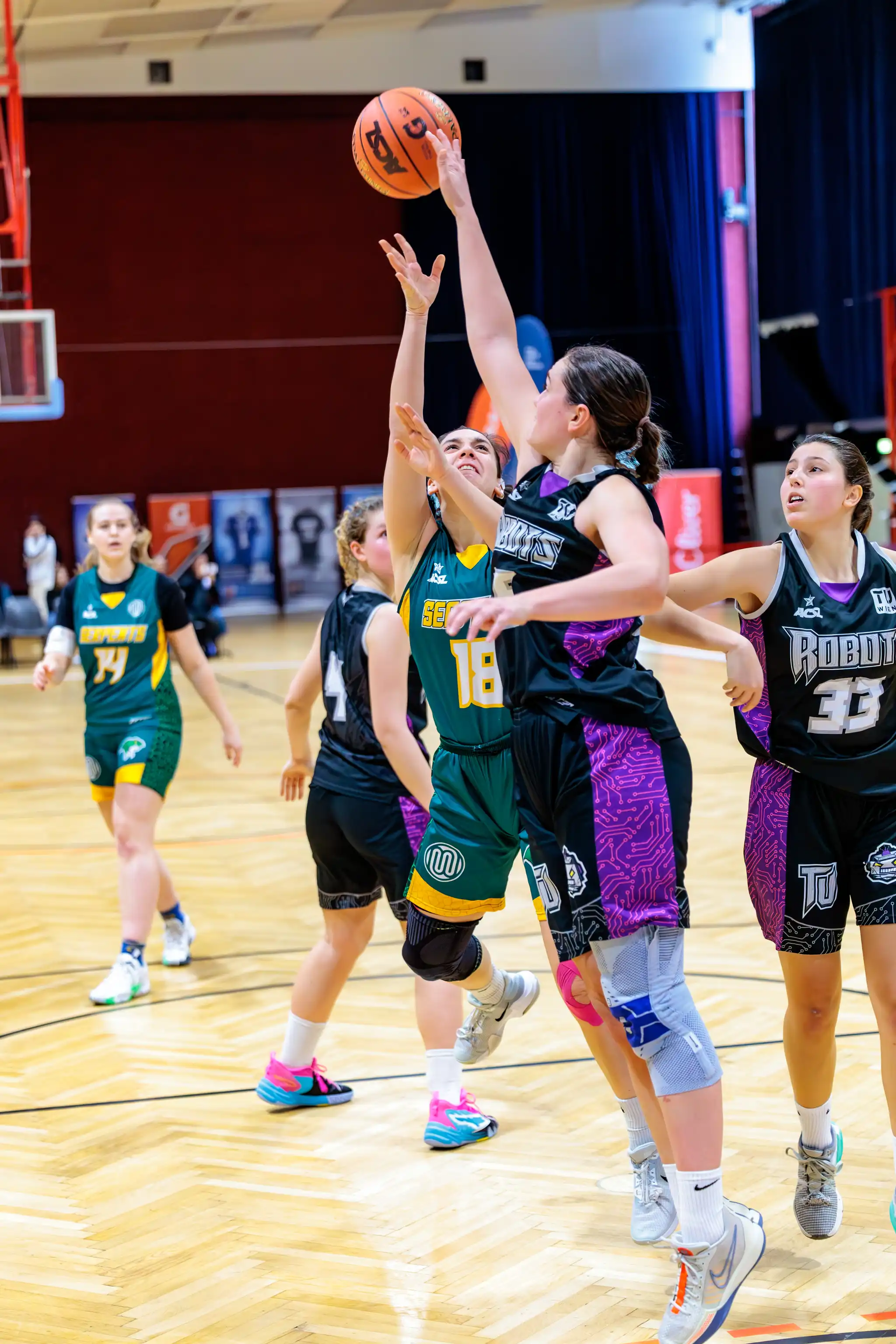 Frauen-Basketballspiel: Eine Spielerin in grün-gelbem Trikot mit der Nummer 18 wirft den Ball, während zwei Gegnerinnen in schwarz-lila Trikots versuchen, sie zu blocken. Andere Spielerinnen sind im Hintergrund auf dem Hallenspielfeld
