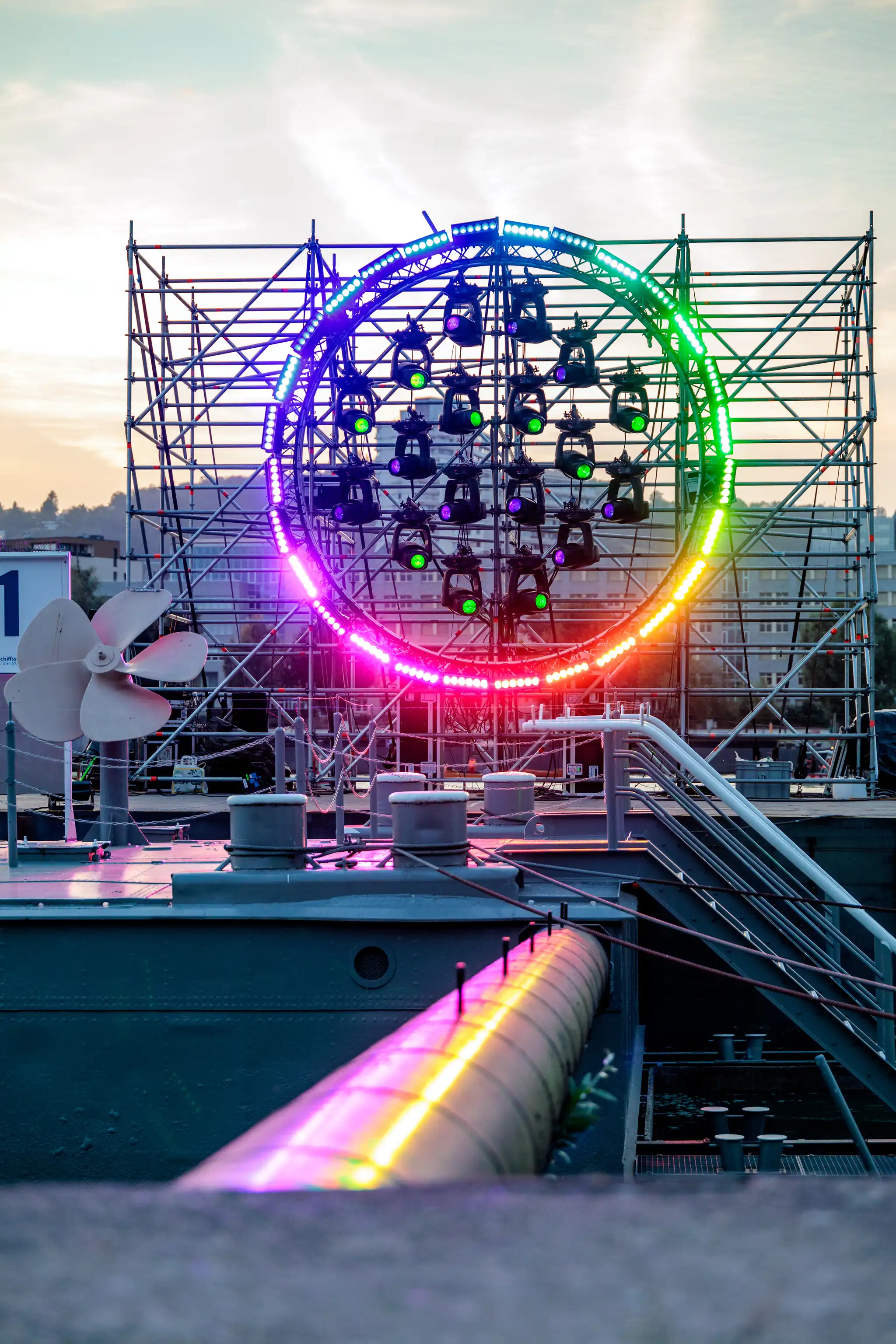 Farbenfroh leuchtende kreisförmige Lichtinstallation mit LEDs und Scheinwerfern auf einer Gerüstkonstruktion bei Dämmerung. Im Vordergrund sind Details eines Schiffes oder Docks wie ein Propeller, Rohre und Poller sichtbar