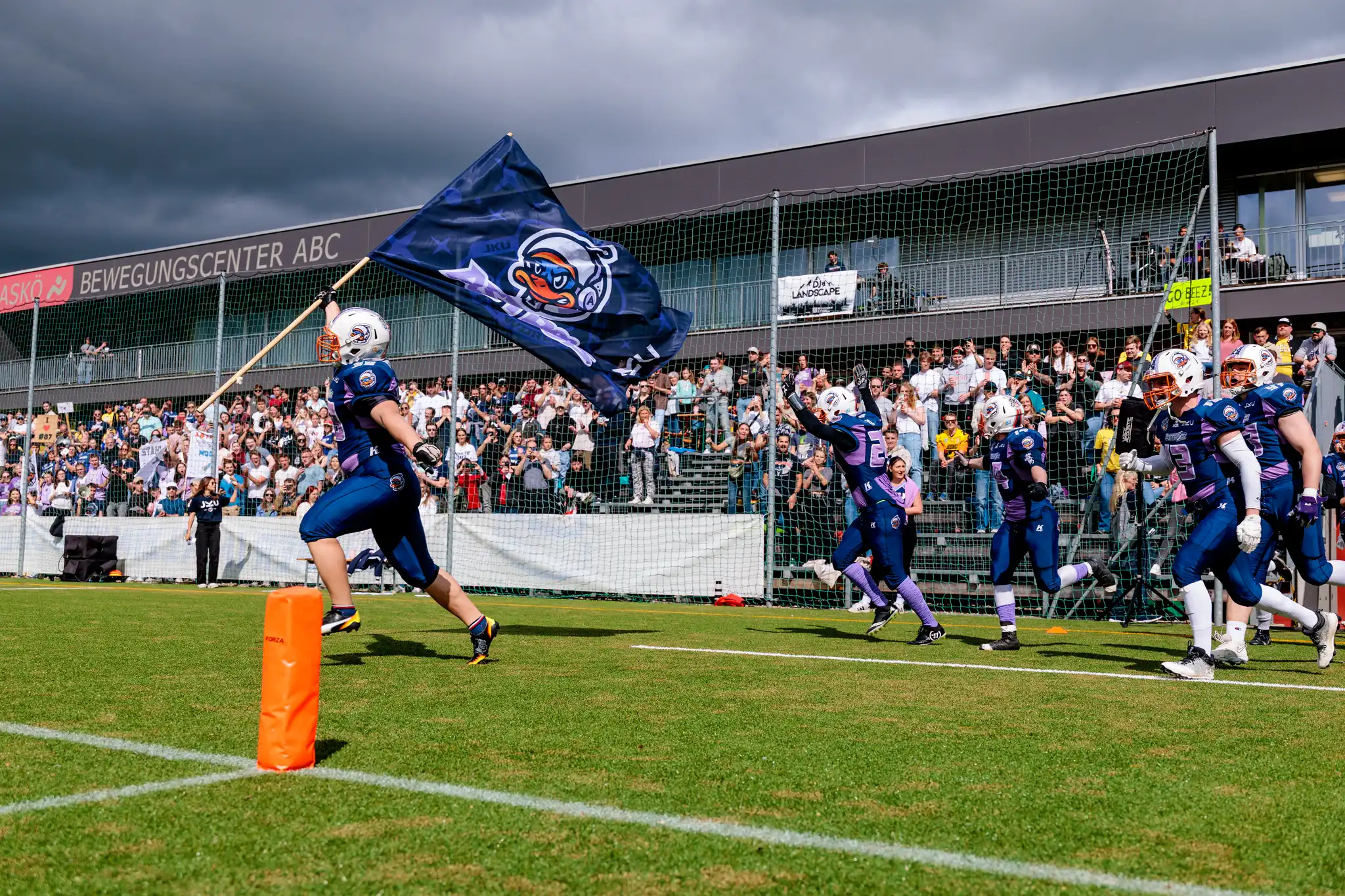 Die JKU Astros betreten das Spielfeld, angeführt von einem Spieler mit einer Teamflagge mit Enten-Logo