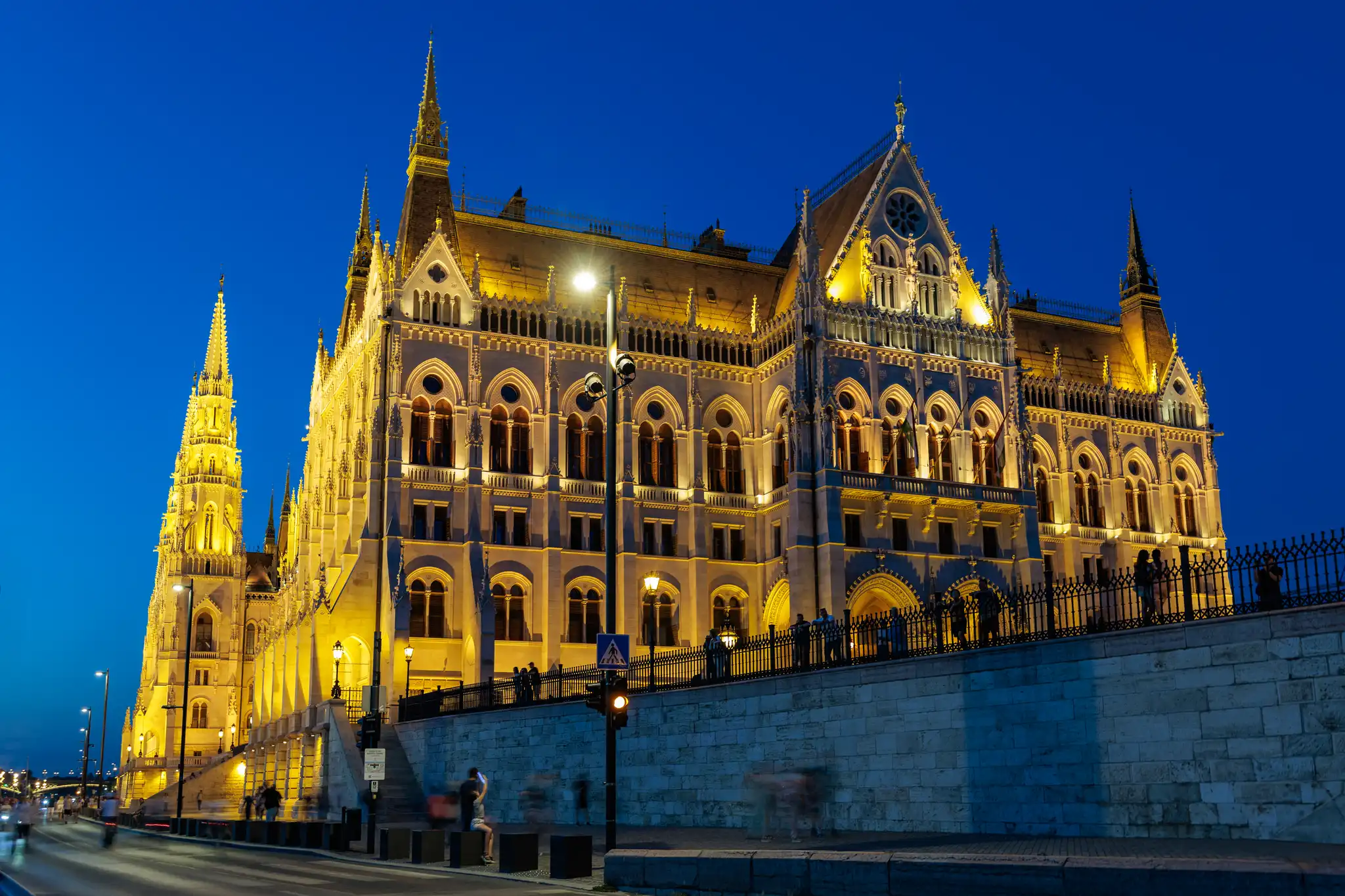 Das beleuchtete Ungarische Parlament (Országház) in Budapest bei Einbruch der Dunkelheit, mit seiner neugotischen Architektur am Donauufer und verschwommenen Menschen.