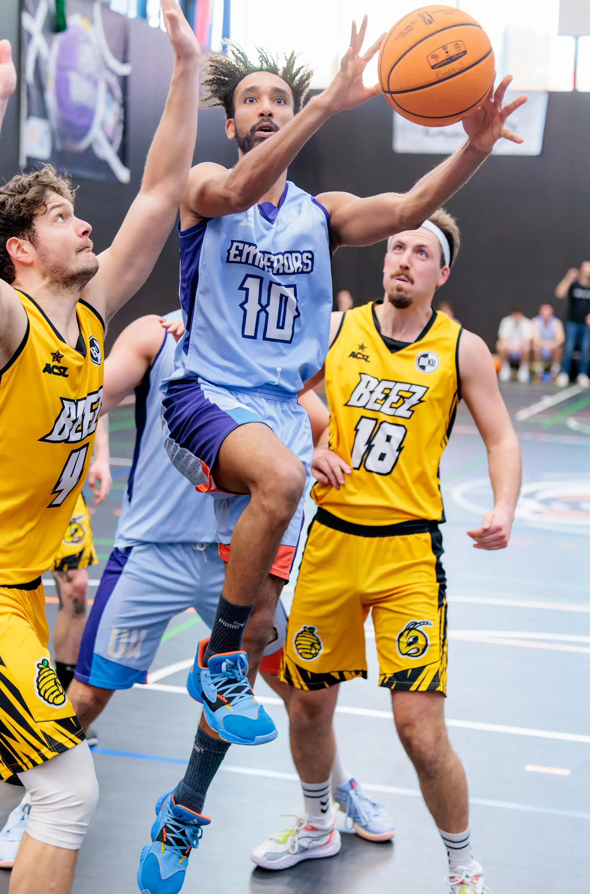 Ein Basketballspieler der Emperors in hellblauem Trikot springt mit dem Ball, während zwei gegnerische Spieler der Beez in gelben Trikots ihn im Spielgeschehen beobachten 