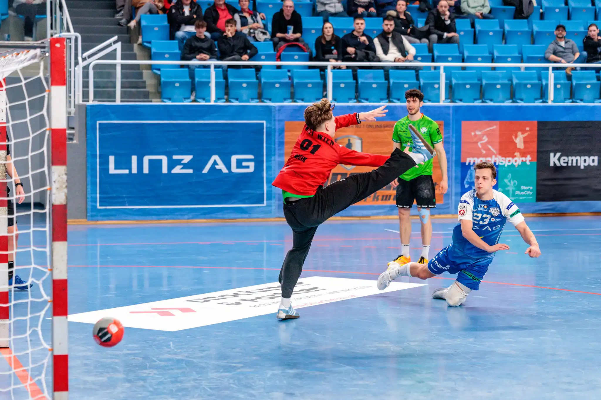Dynamisches Handballspiel: Torwart in Rot pariert Wurf, Spieler in Blau liegt am Boden. Aktion in der Halle