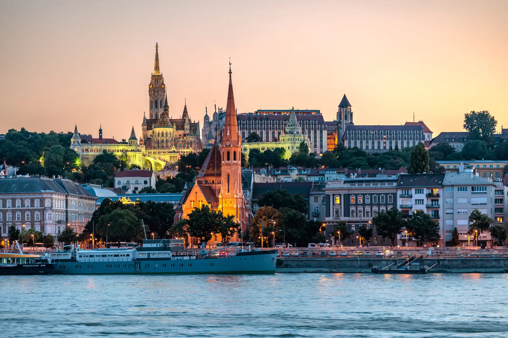 Stadtansicht von Budapest bei Sonnenuntergang mit der beleuchteten Matthiaskirche, Fischerbastei und dem Budaer Burgviertel am Donauufer.