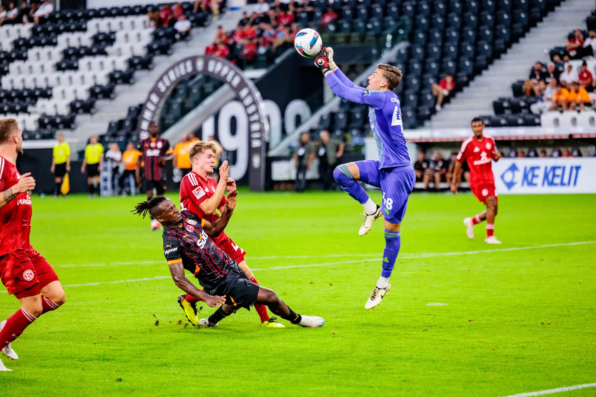 Ein dynamisches Fußballspiel zeigt einen Torhüter, der den Ball in der Luft abfängt, während zwei Feldspieler auf dem Rasen in einen Zweikampf verwickelt sind