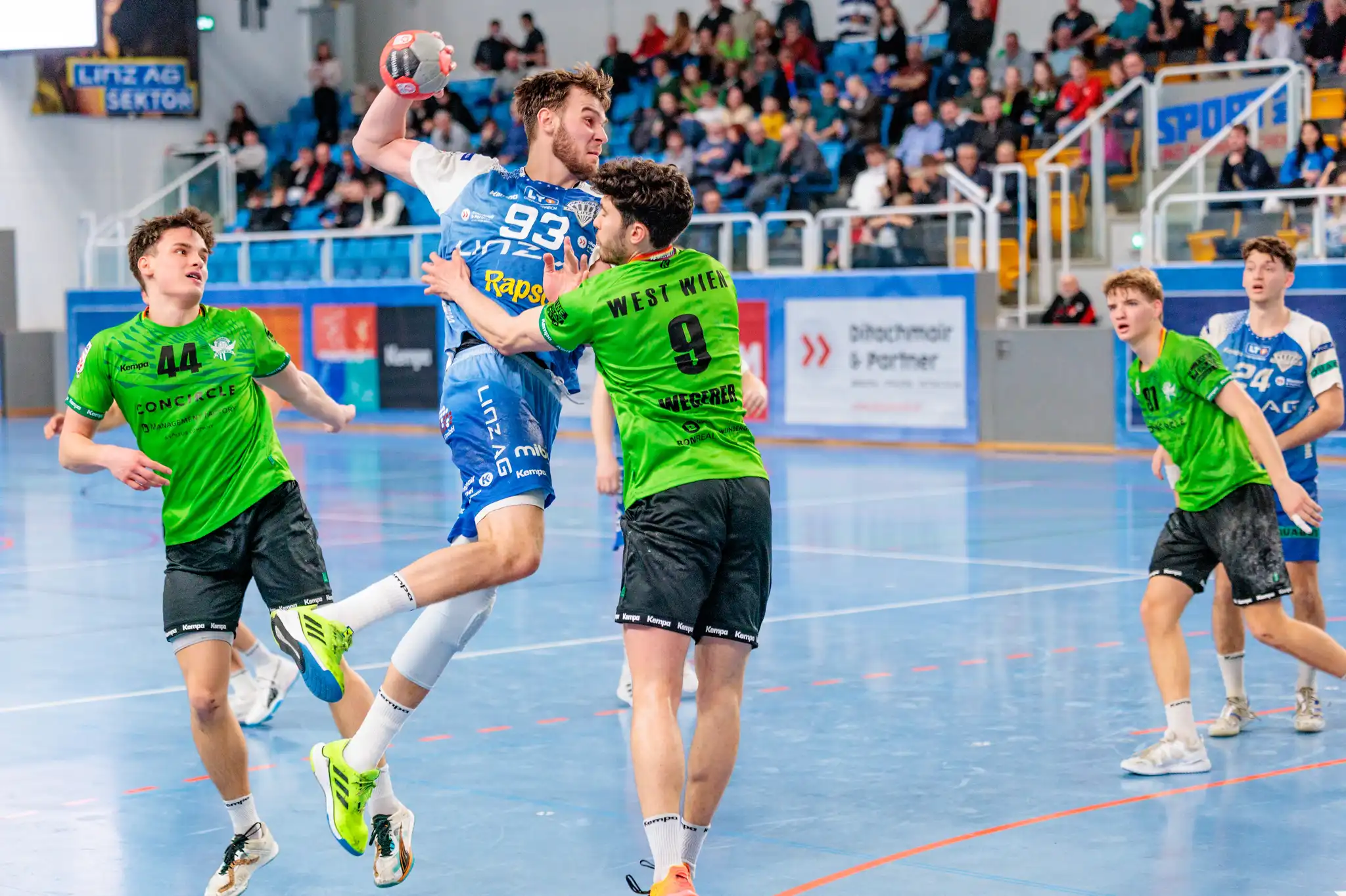 Dynamisches Handballspiel mit einem Spieler im blauen Trikot (Nr. 93) im Sprungwurf, verteidigt von Spielern in grünen Trikots, auf einem blauen Spielfeld