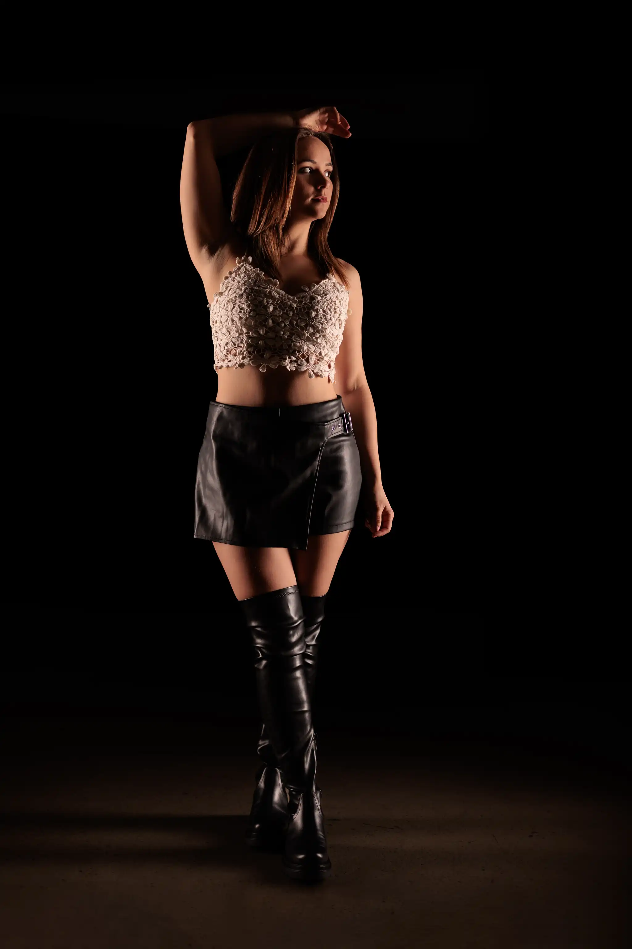 Weibliches Modell in hellem Häkel-Crop-Top, schwarzem Leder-Minirock und Overknee-Stiefeln, posiert auf dunklem Hintergrund mit dramatischer Low-Key-Beleuchtung. 