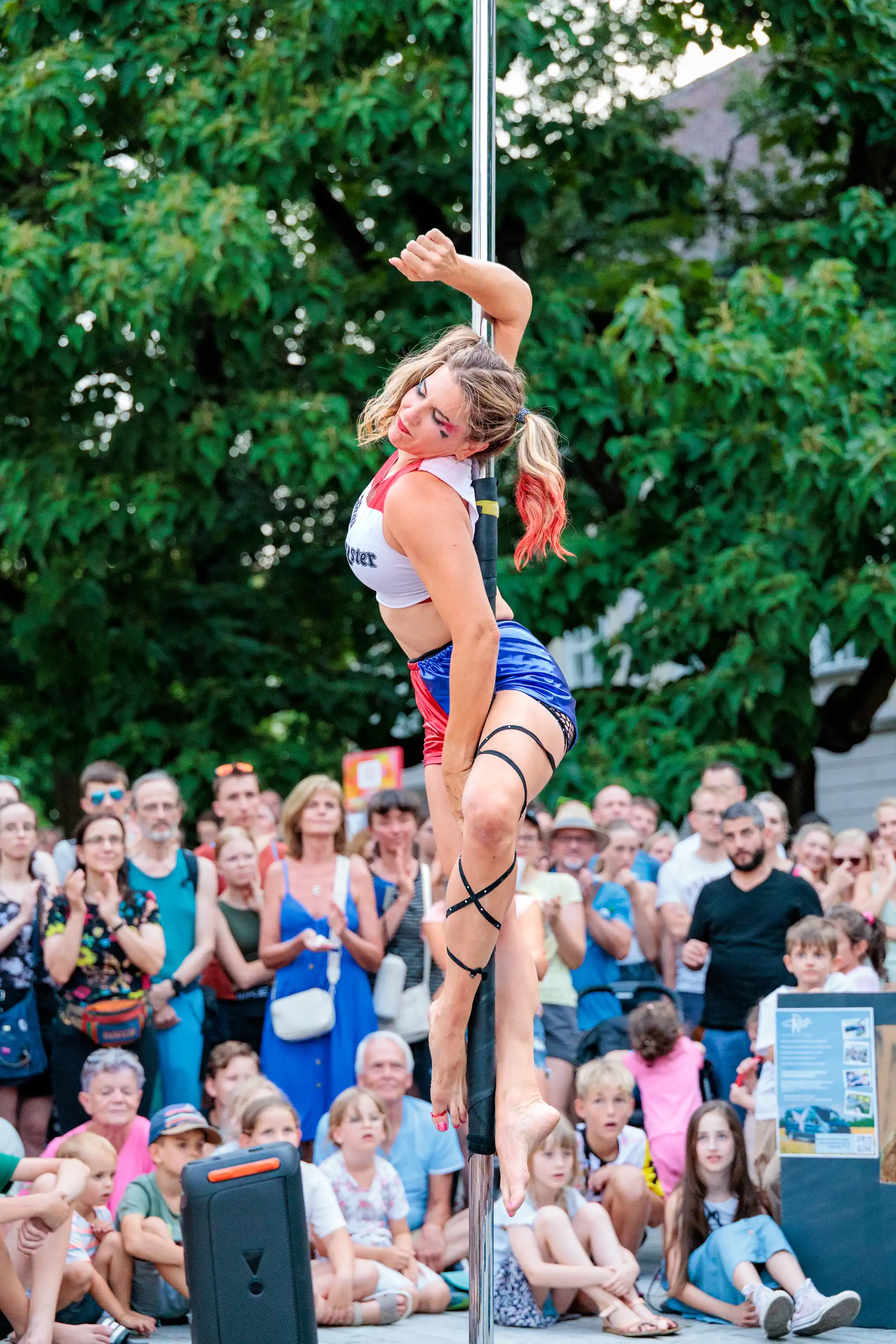 Poledancerin in akrobatischer Pose an der Stange bei einer Outdoor-Show vor einem Publikum mit grünen Bäumen im Hintergrund