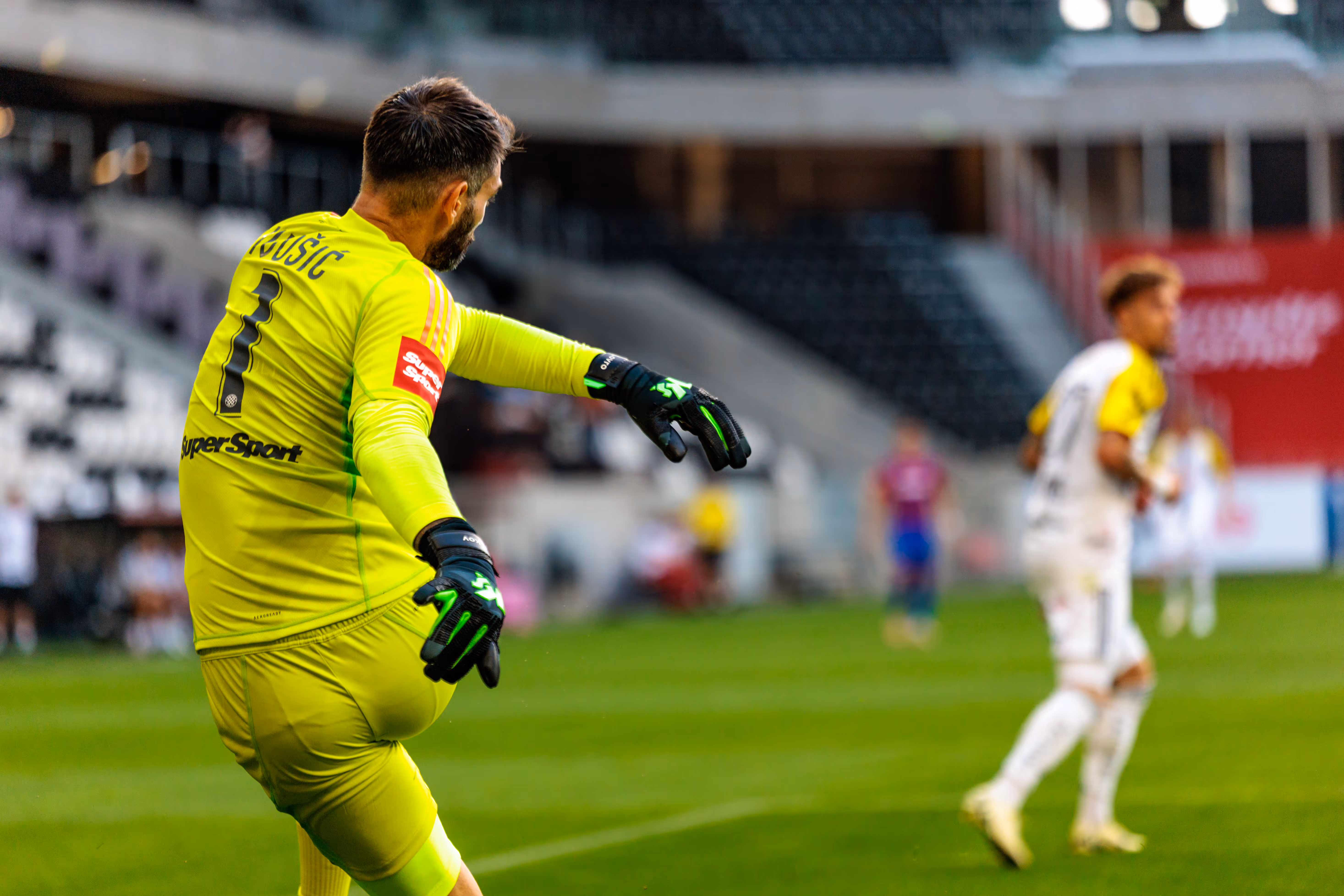 Ein Fußballtorhüter in leuchtend gelb-grünem Trikot mit der Nummer 1 und dem Namen Družić ist während eines Spiels in einem Fußballstadion zu sehen - 2025/07/18