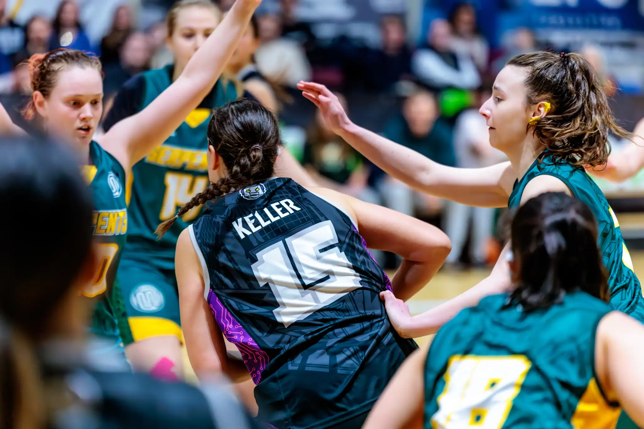 Dynamisches Frauen-Basketballspiel, das eine Spielerin im schwarzen Trikot mit KELLER und der Nummer 15 auf dem Rücken zeigt, umgeben von gegnerischen Basketballspielerinnen in grünen Trikots auf dem Hallenfeld
