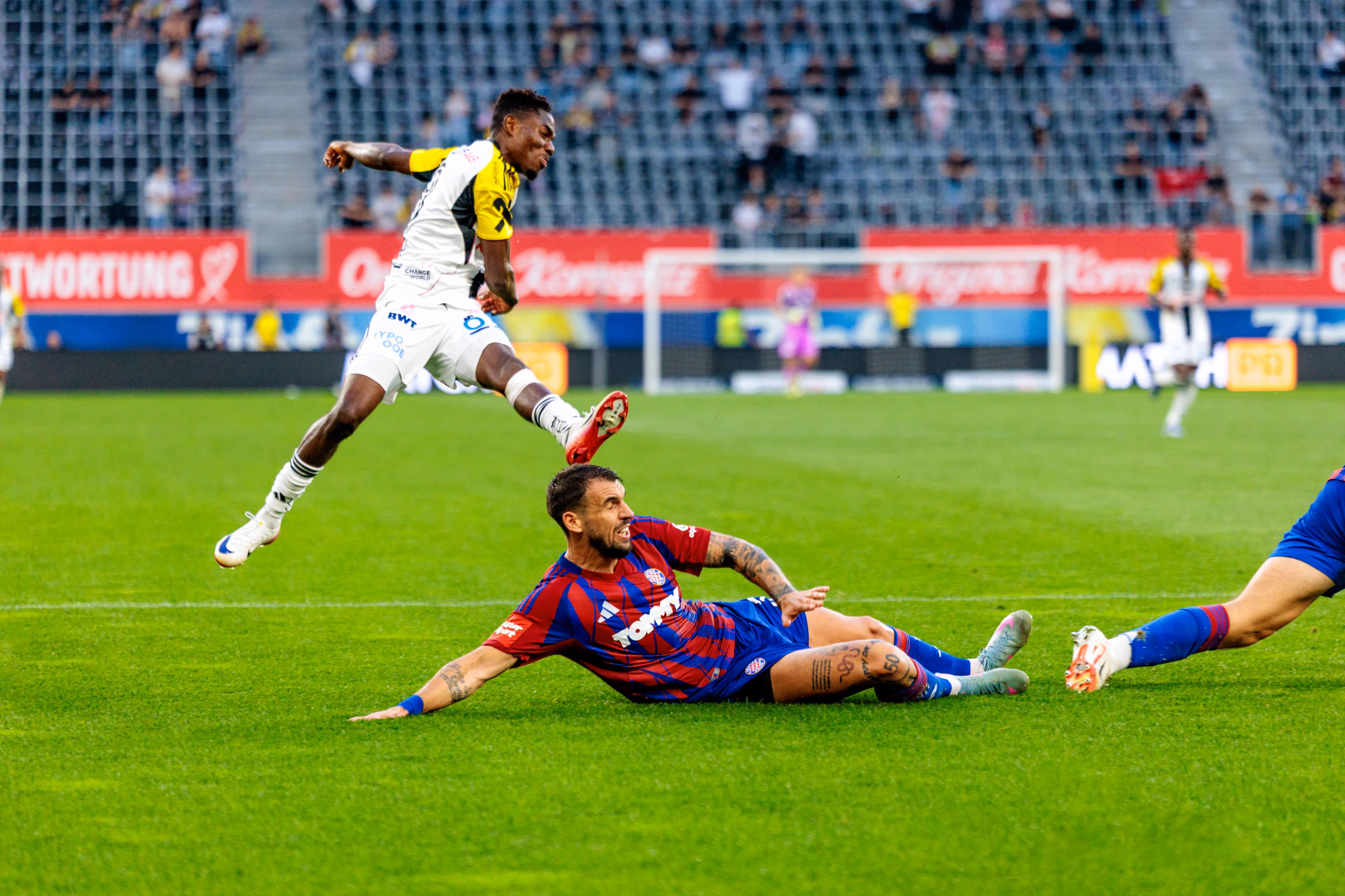 Ein Fußballspieler des LASK springt nach einem Schuss über einen über den Boden rutschenden Gegenspieler