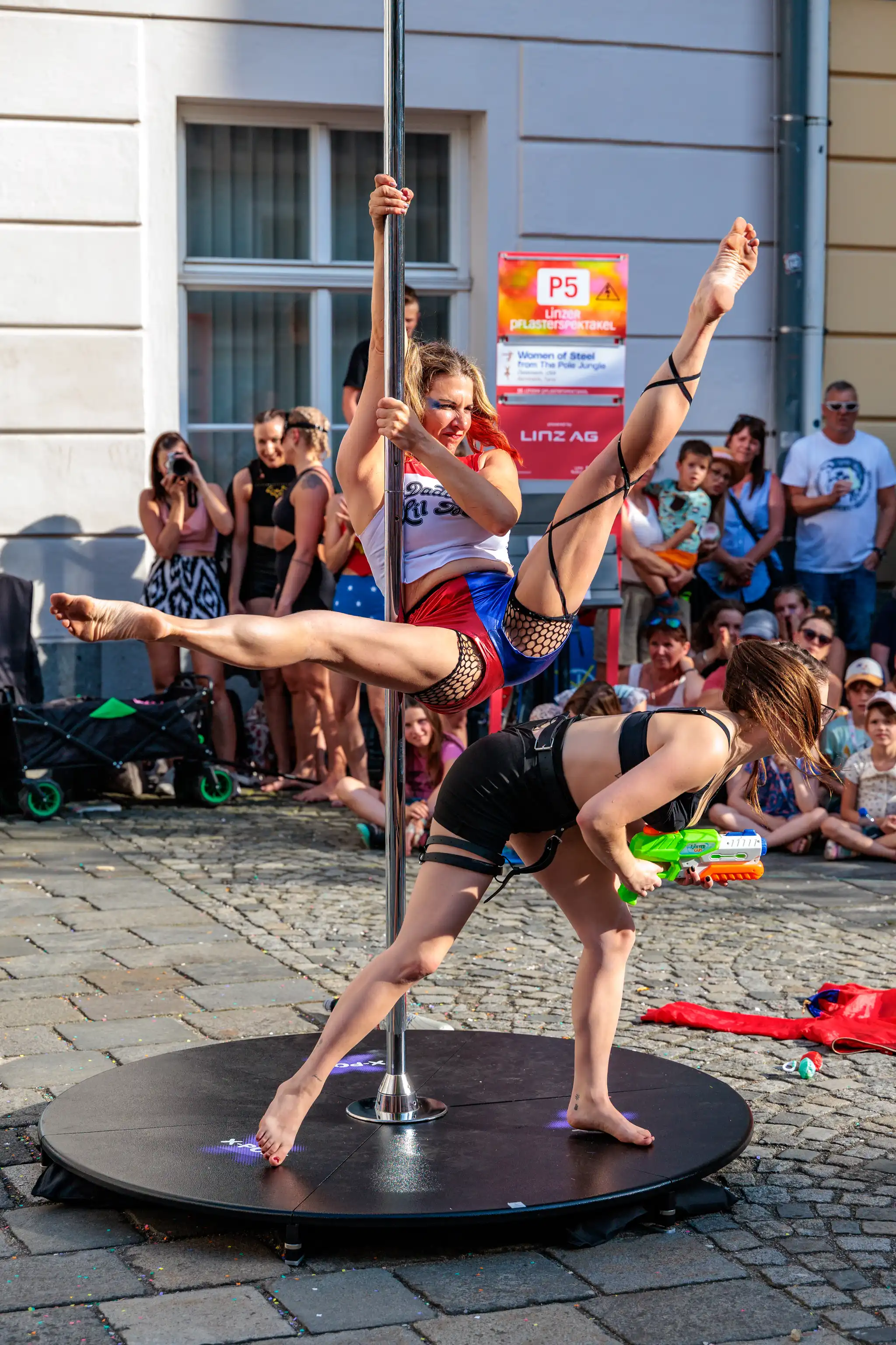 Outdoor Pole Dance Performance auf dem Linzer Pflasterspektakel: Zwei Frauen führen Akrobatik am Mast vor. Eine Sportlerin in rot-blauer Verkleidung zeigt einen Spagat, während eine andere Frau in Schwarz mit einer Wasserpistole posiert. Im Hintergrund beobachtet Publikum die Szene