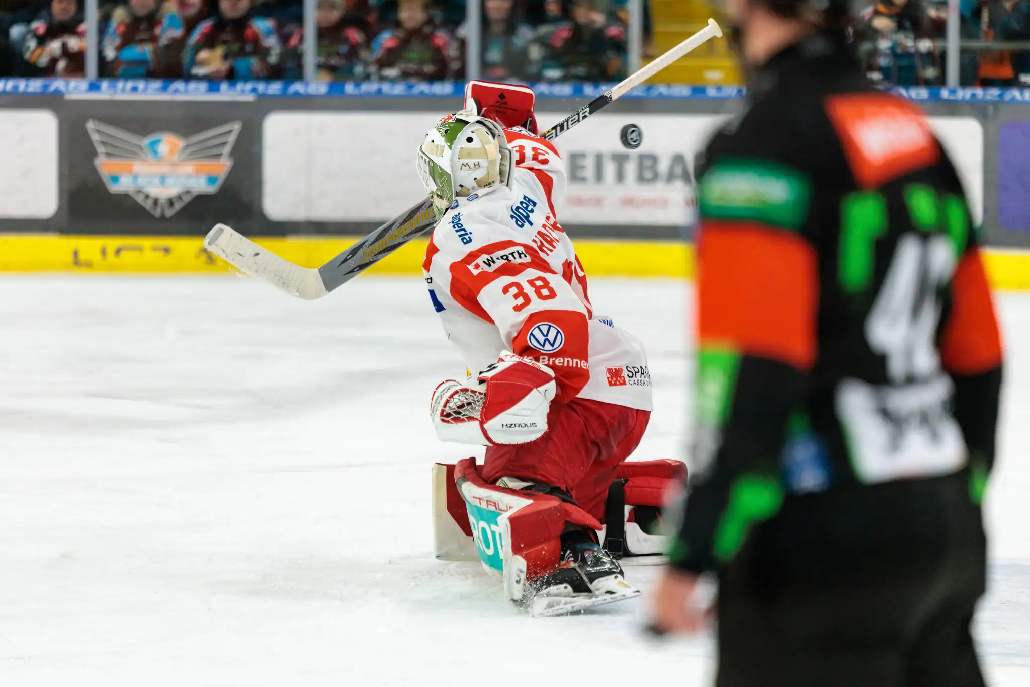 Eishockey-Torwart Nummer 38 in rot-weißer Ausrüstung pariert einen Puck auf dem Eis, im Hintergrund die Linz AG Eisarena