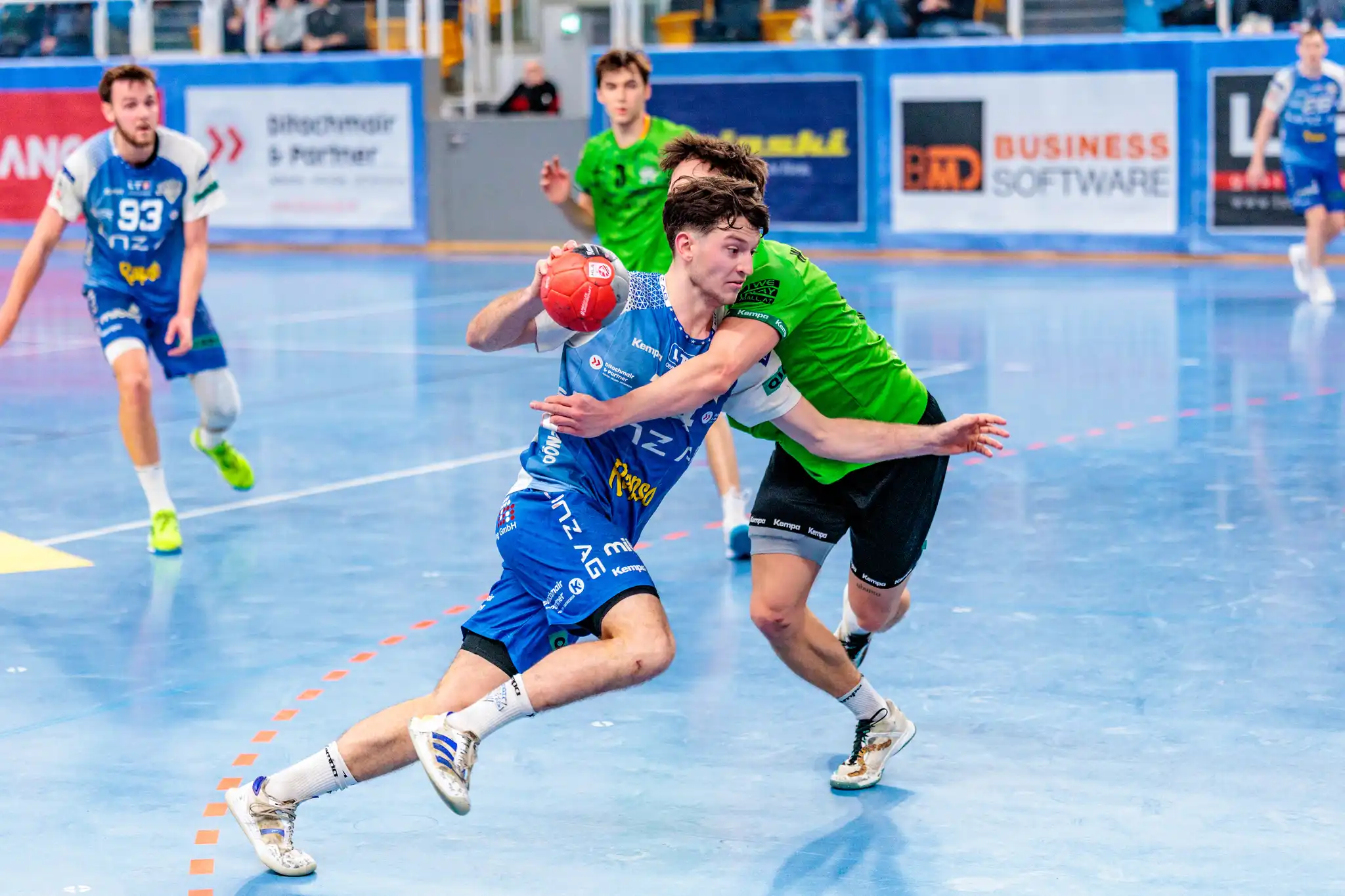 Dynamisches Handballspiel: Spieler im blauen Trikot mit rotem Ball wird von Gegenspieler im grünen Trikot verteidigt