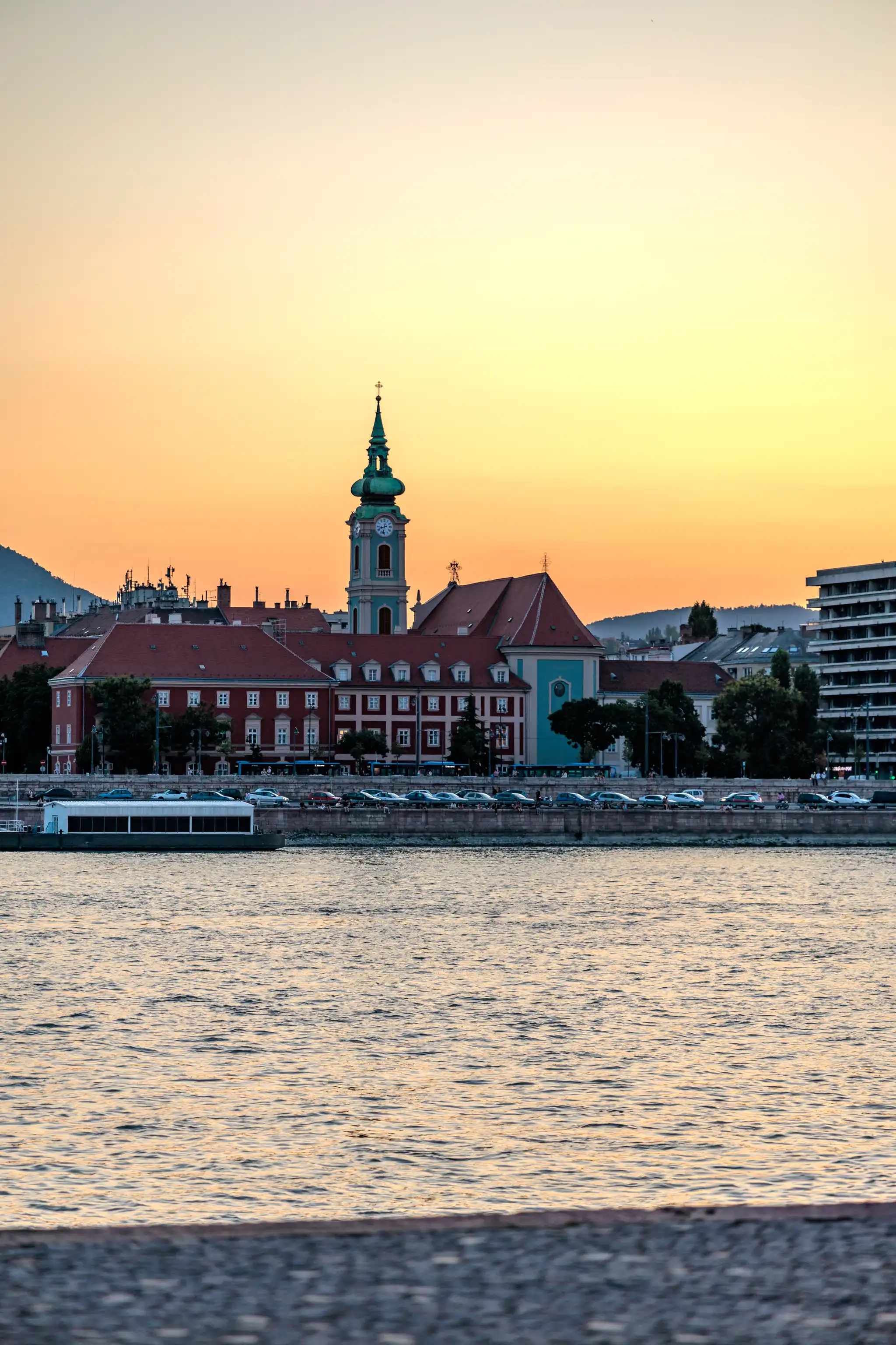 Abendstimmung über der Donau in Budapest mit der prominenten St.-Anna-Kirche, historischen Gebäuden und einem Flussboot am Ufer.