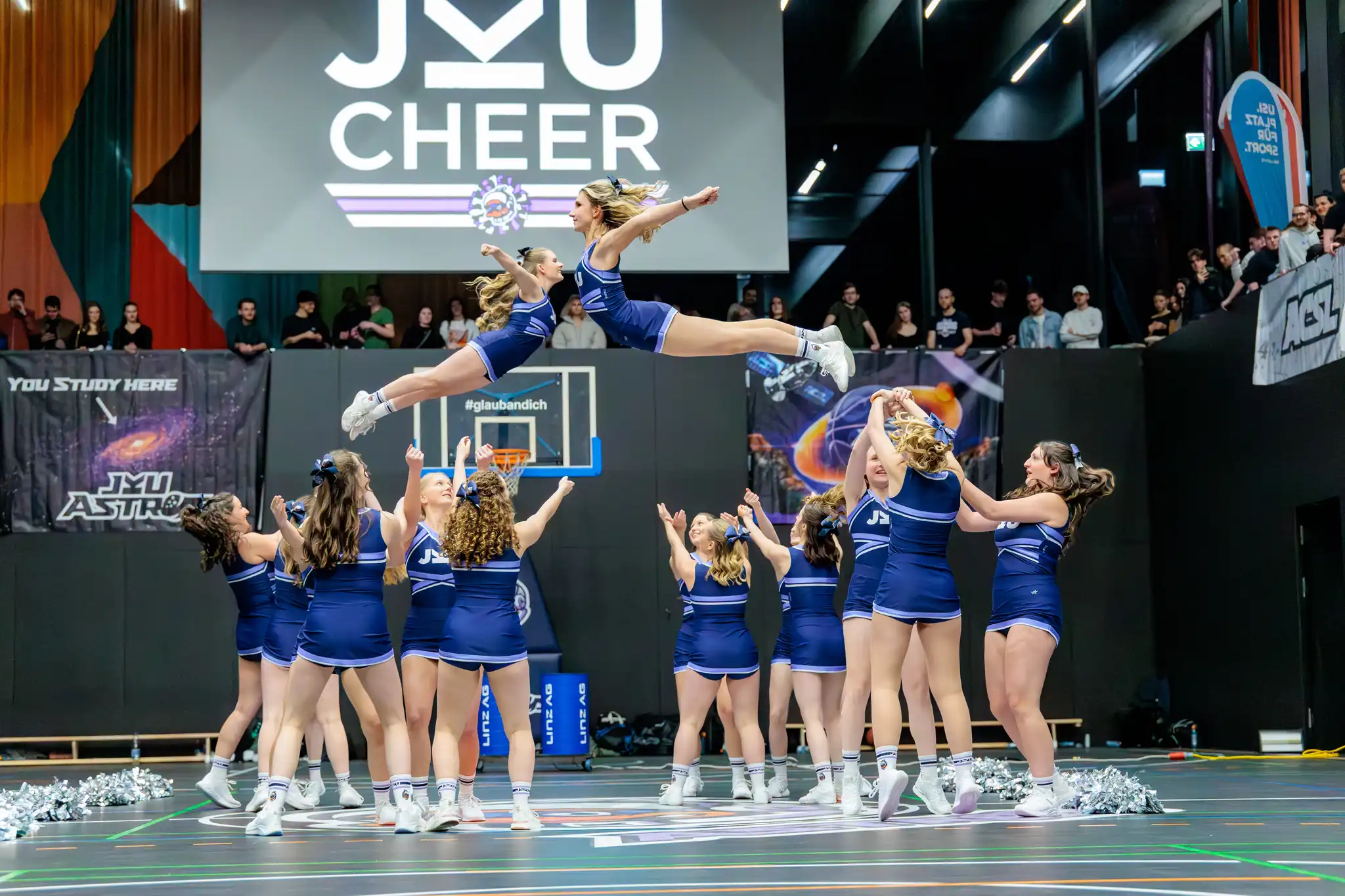 Cheerleader-Team in blauen Uniformen bei einem dynamischen Stunt in einer Sporthalle. Zwei Cheerleaderinnen schweben horizontal in der Luft, gestützt von den Teammitgliedern am Boden. Im Hintergrund ist das JKU CHEER Logo zu sehen