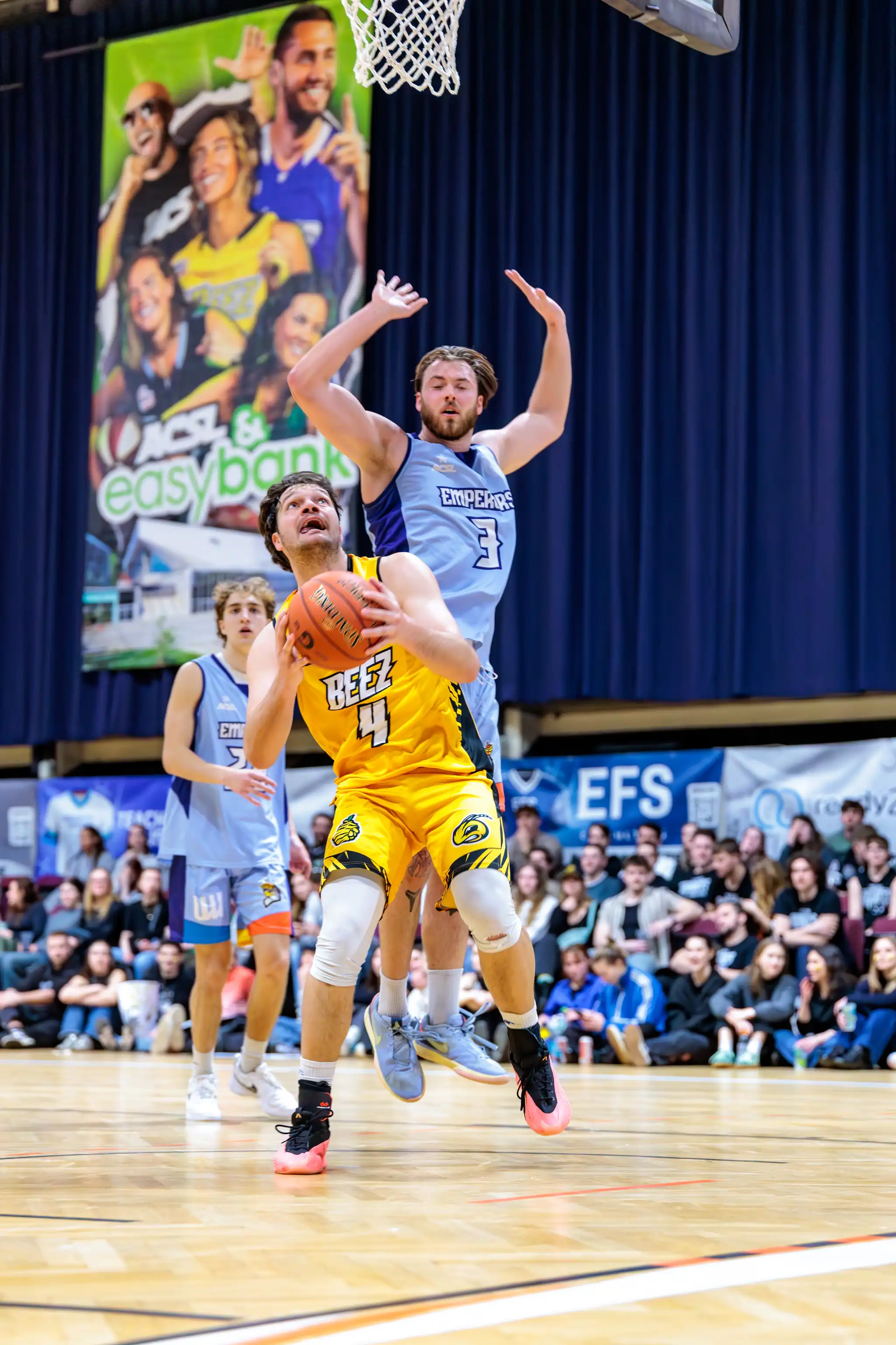 Zwei Basketballspieler im Sprung während eines Spiels: Ein Spieler im gelben Trikot mit Ball wird von einem Gegenspieler in Hellblau verteidigt. Im Hintergrund sind Zuschauer und Werbebanner auf einem Indoor-Basketballfeld zu sehen 