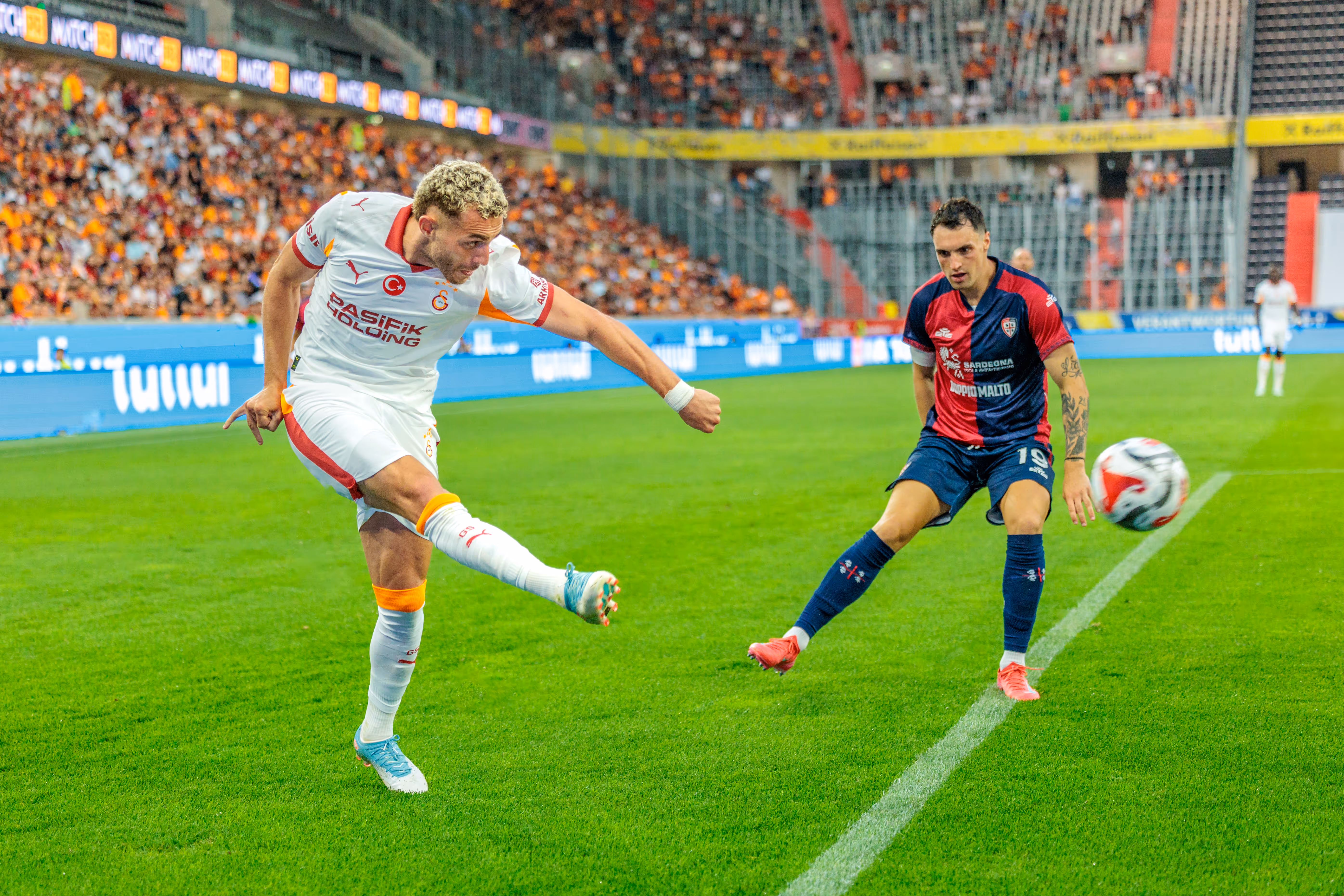 Ein Galatasaray-Fußballspieler in weißem Trikot schießt den Ball in einem Stadion, beobachtet von einem Cagliari-Spieler