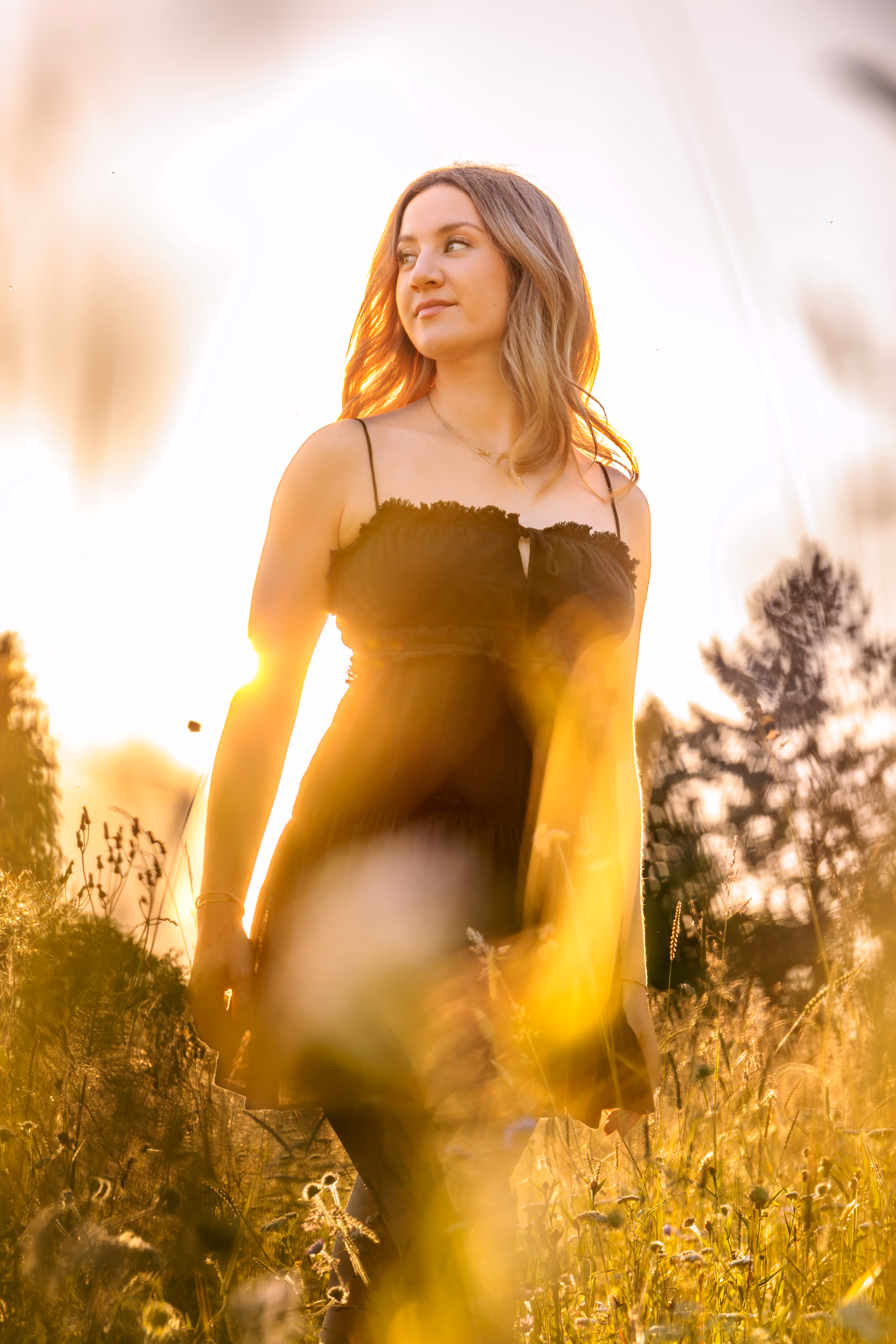 Eine junge Frau im dunklen Kleid steht bei goldenem Gegenlicht in einer sonnigen Sommerwiese