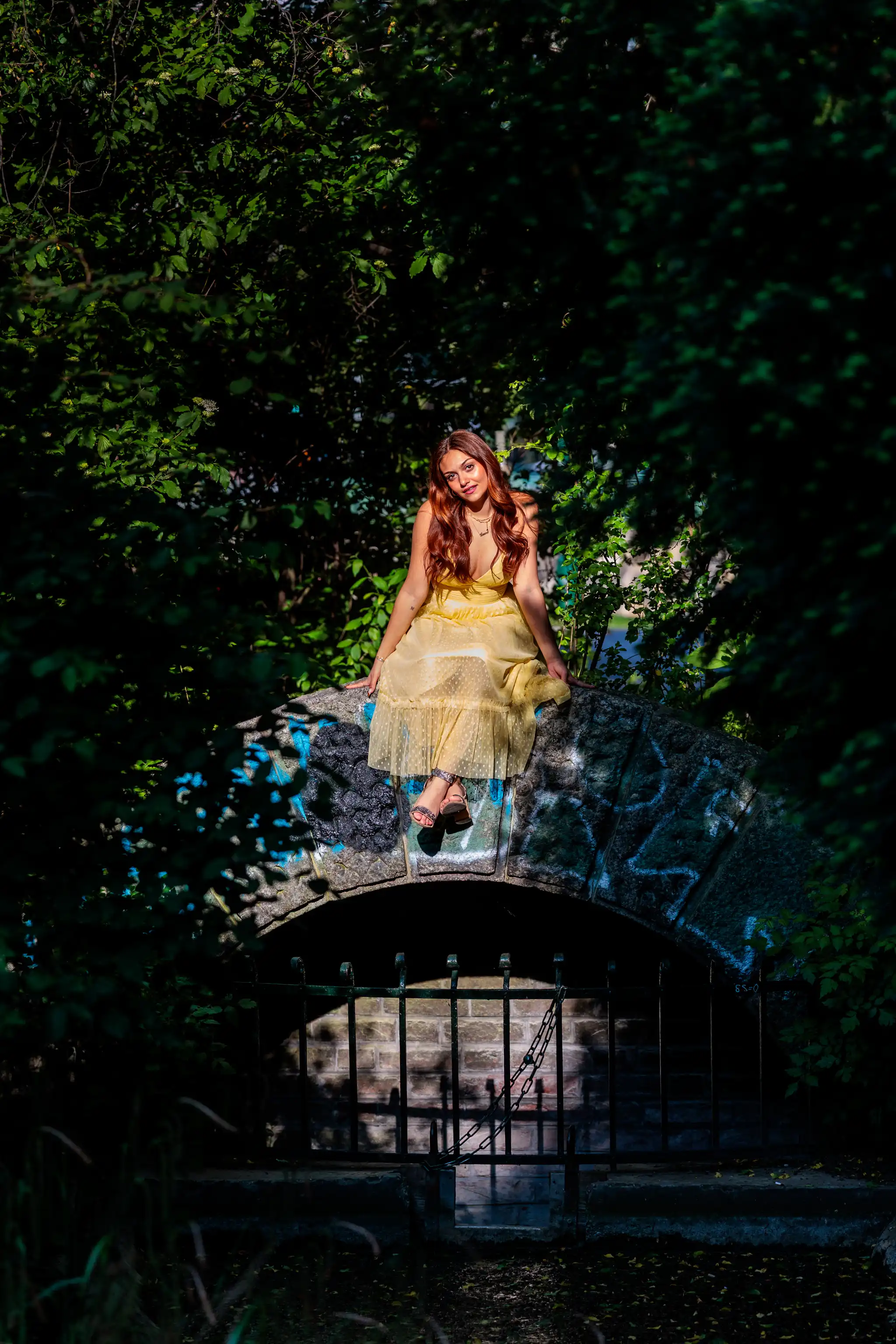 Junge Frau im gelben Sommerkleid sitzt im Sonnenlicht auf einer alten Steinbogenbrücke mit Graffiti, umrahmt von dichtem Grün. 