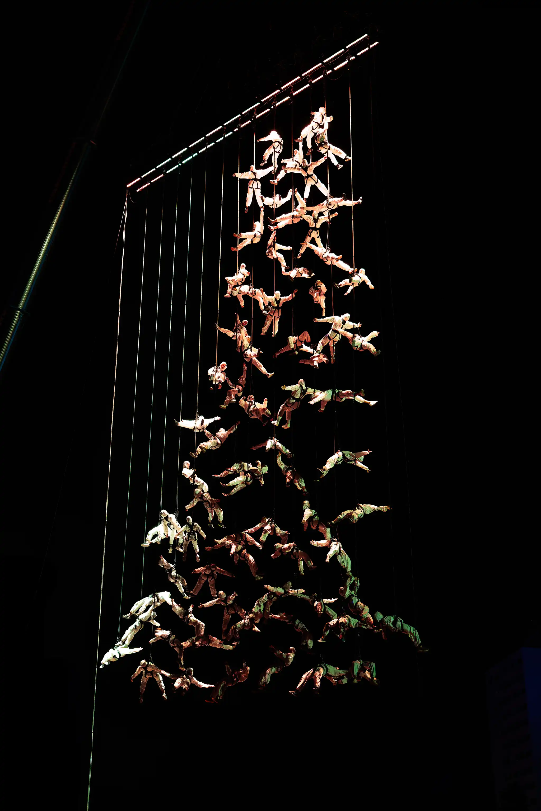 Luftakrobatik-Performance während der Linzer Klangwolke 2024 mit vielen Artisten in hellen Anzügen, die an Seilen von einer beleuchteten Deckenkonstruktion herabhängen und eine schwebende Formation vor dem nächtlichen Himmel bilden