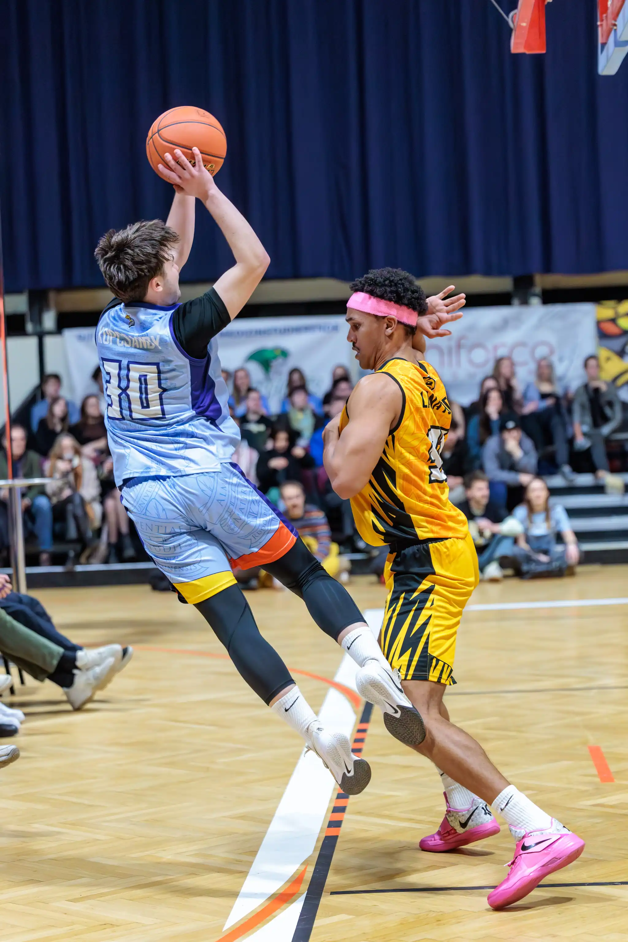 Dynamisches Basketballspiel: Ein Spieler im hellblauen Trikot setzt zu einem Sprungwurf an, neben einem Verteidiger mit pinkem Stirnband im gelben Trikot. Szene in einer Sporthalle 