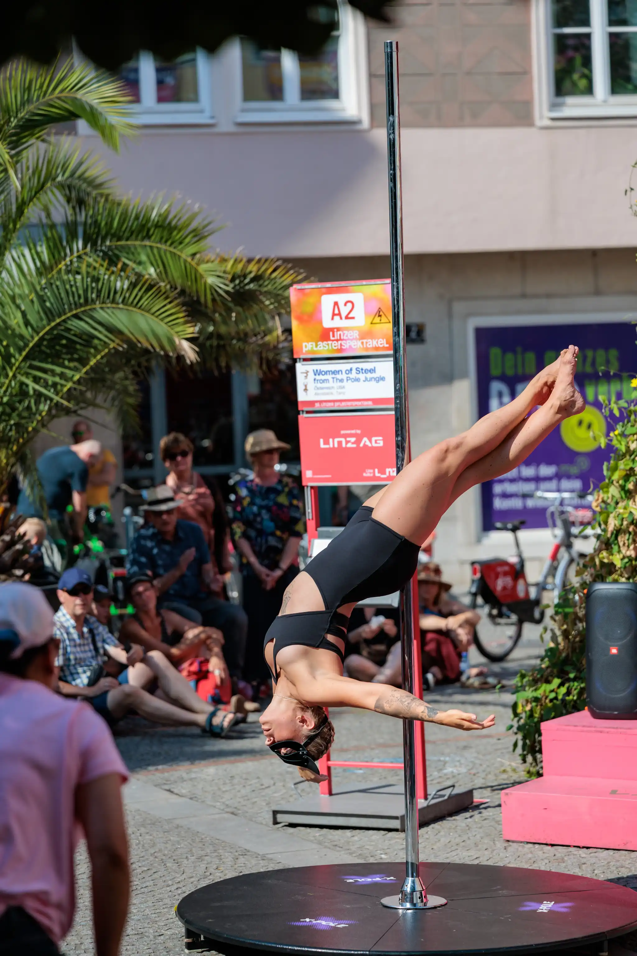Eine Poledance-Künstlerin in schwarzem Bodysuit und dunkler Maske führt kopfüber eine akrobatische Figur an einer X-Pole Stange aus, umgeben von Zuschauern beim Linzer Pflasterspektakel. Im Hintergrund sind ein Palmenbaum, Gebäude und Schilder mit "Linzer Pflasterspektakel" und "Women of Steel from The Pole Jungle" zu sehen