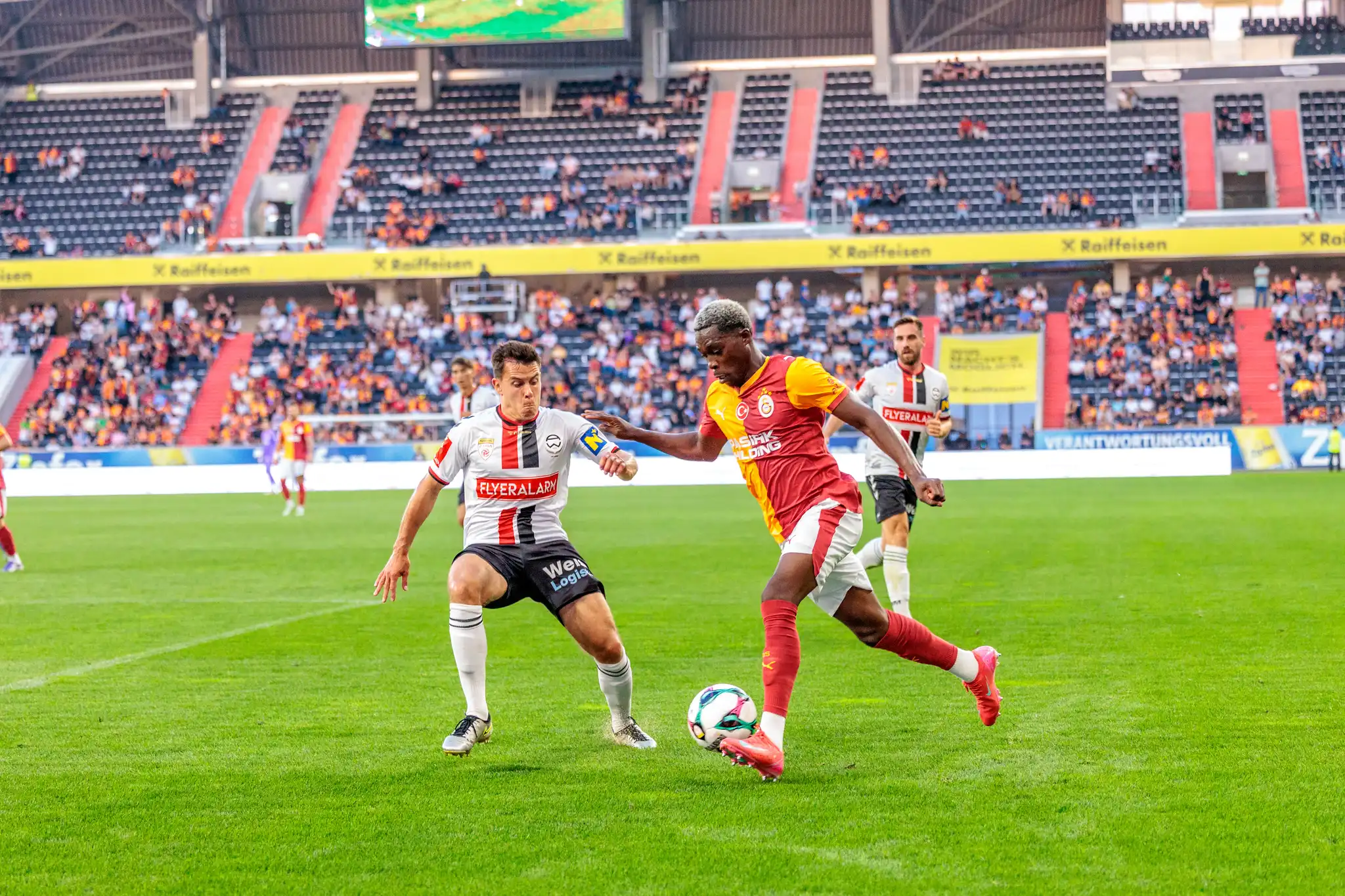 Zwei Fußballspieler, ein Admira Wacker und ein Galatasaray Spieler, duellieren sich im Spiel um den Ball in der Raiffeisen Arena vor Zuschauern 