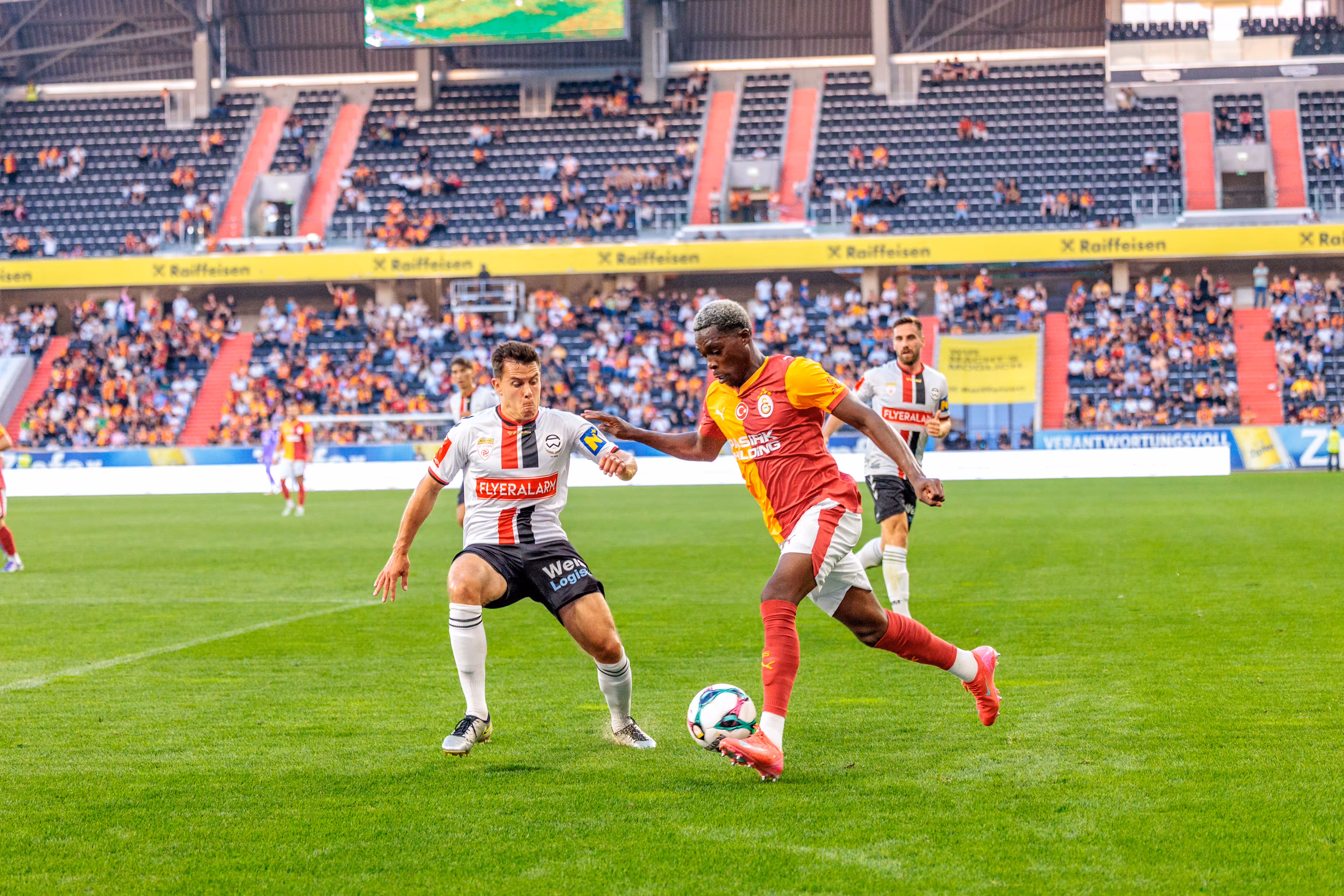 Zwei Fußballspieler, ein Admira Wacker und ein Galatasaray Spieler, duellieren sich im Spiel um den Ball in der Raiffeisen Arena vor Zuschauern