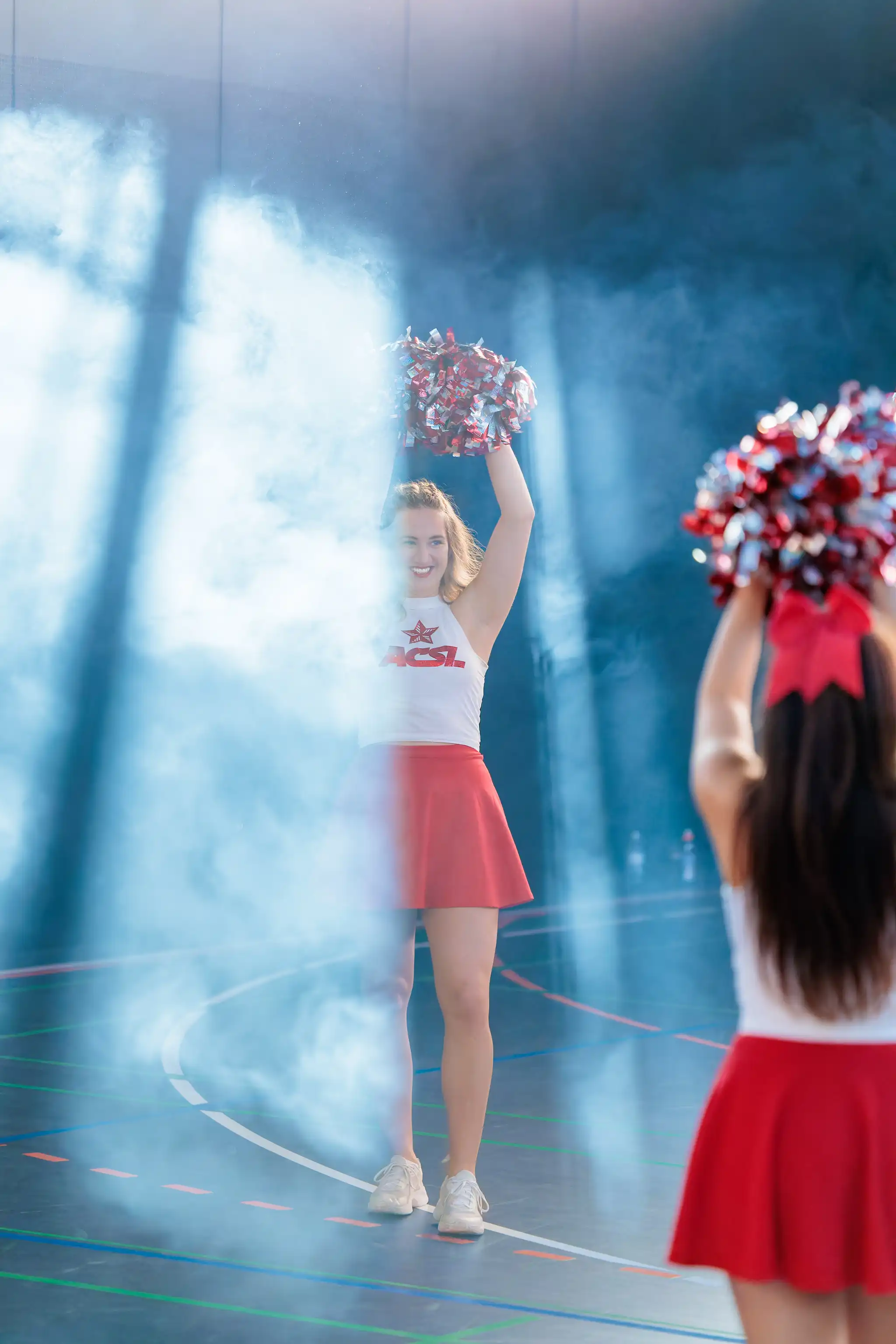 Eine lächelnde Cheerleaderin in rot-weißem Outfit hält ihre Pom-Poms in einer rauchigen Halle hoch, während eine zweite bei einem ACSL Basketball-Halbfinale im Vordergrund sichtbar ist