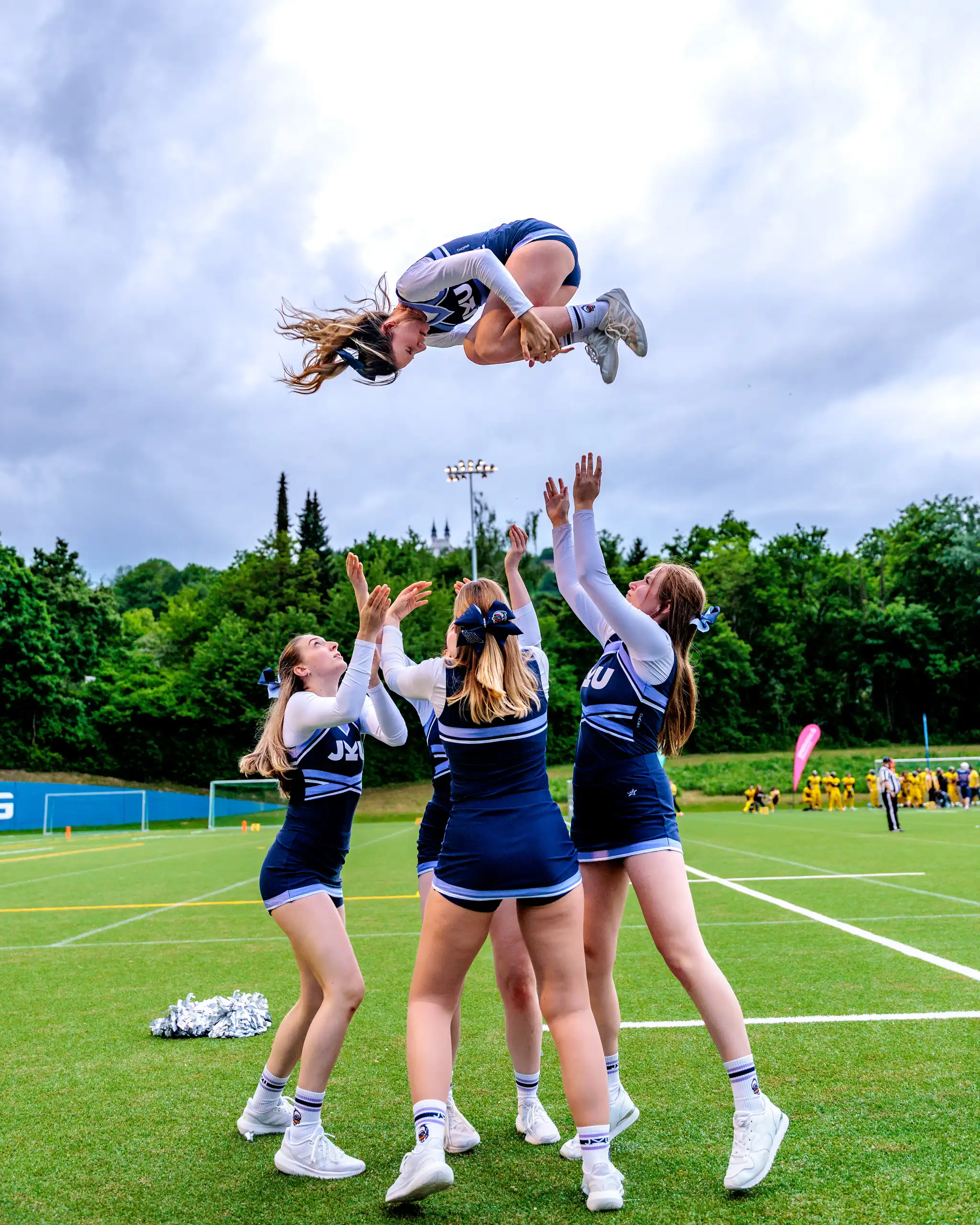 Eine Cheerleaderin vollführt einen akrobatischen Salto hoch in der Luft, umgeben von ihren Teamkolleginnen, die bereit sind, sie zu fangen