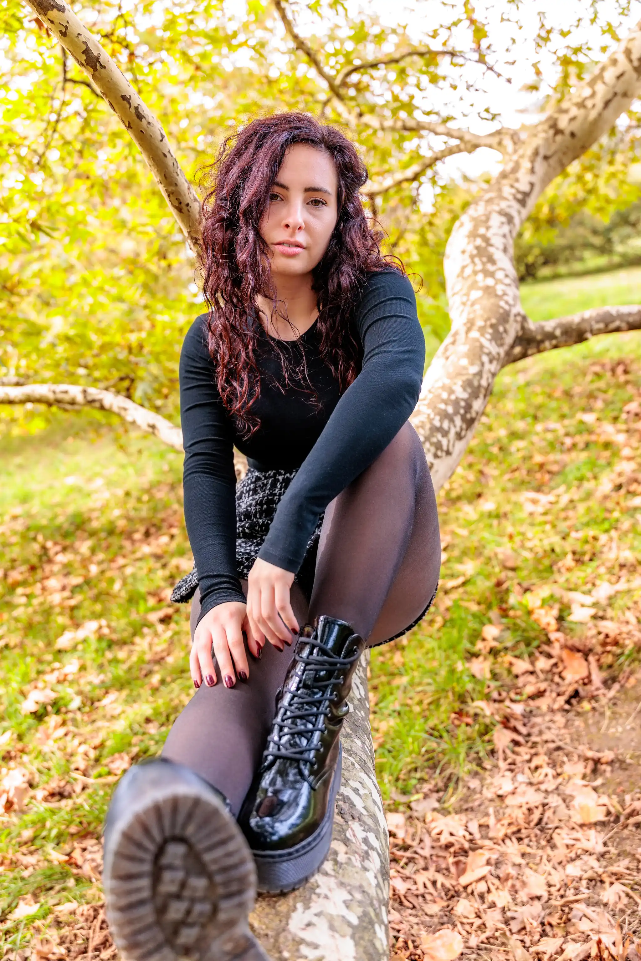Junge Frau mit langen, dunkelroten Locken in schwarzem Oberteil, dunkler Strumpfhose und glänzenden schwarzen Plateau-Stiefeln sitzt auf einem markanten Baumstamm im Herbstlaub im Freien und blickt direkt in die Kamera.